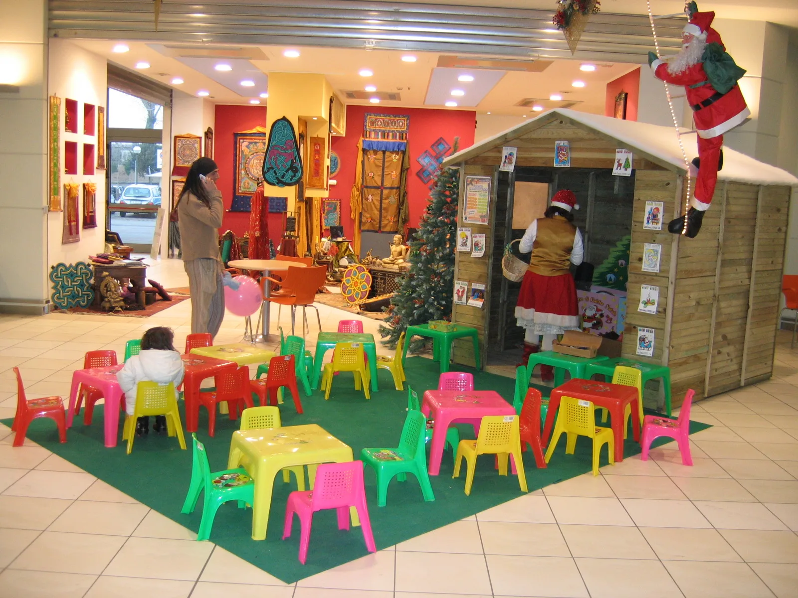 Area bimbi natalizia con la casetta di legno di Babbo Natale, l'elfa che consegna i regali, il tappeto verde con i tavolini per i disegni e Babbo Natale che si arrampica sulla parete