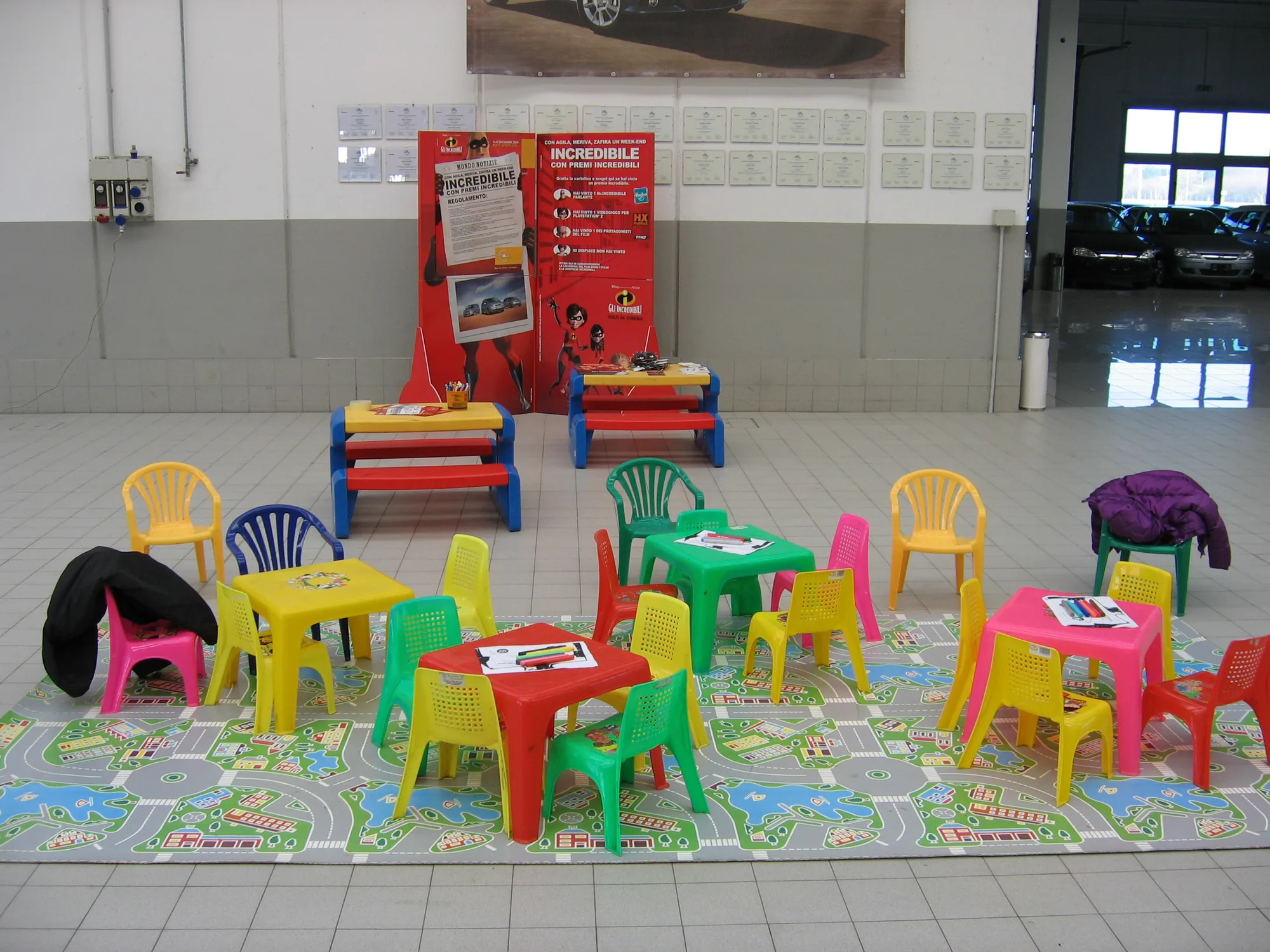 Tappeto gioco con le strade disegnate, tavolini colorati con fogli e pennarelli e panchine da lavoro allestiti come area bimbi dentro una concessionaria