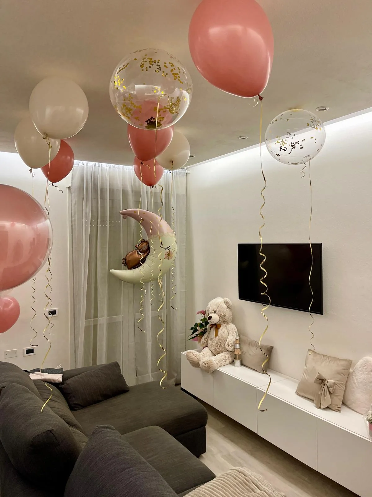Salotto di casa con palloncini a elio rosa, panna e trasparenti con coriandoli oro attaccati al soffitto e un palloncino luna con orsetto al centro