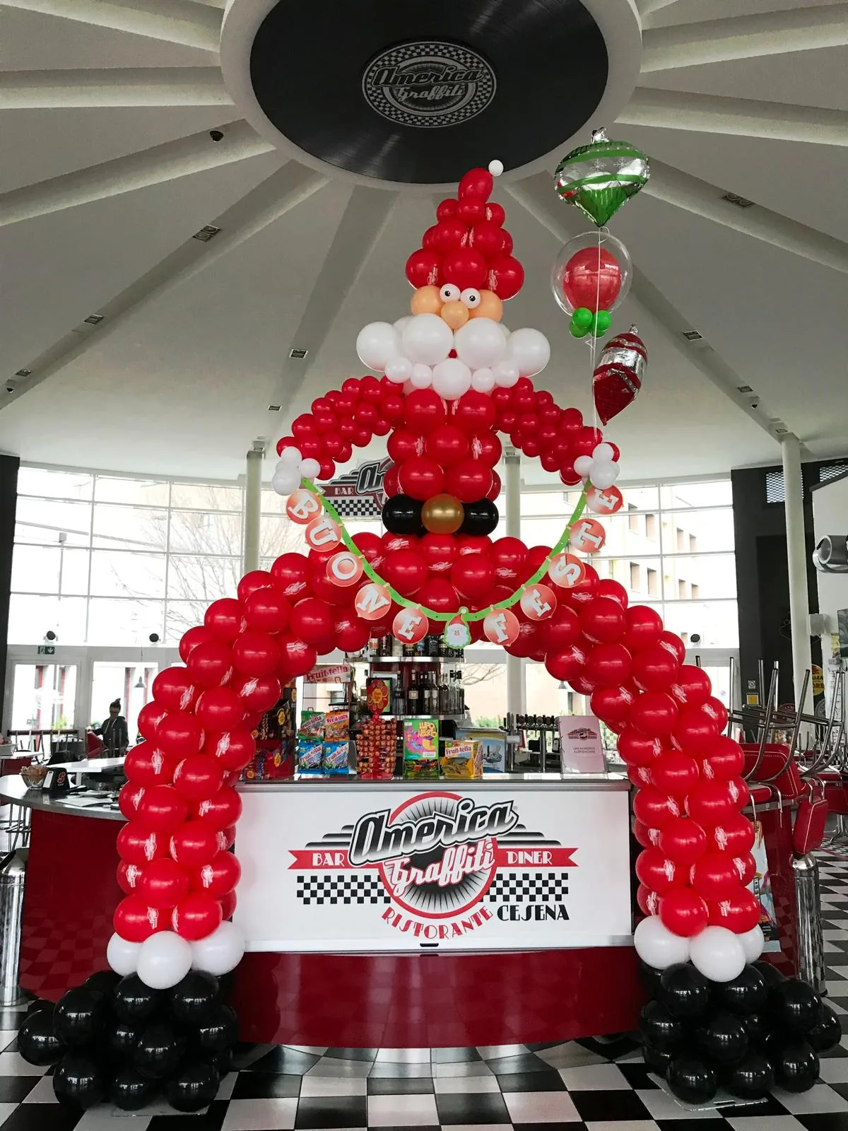 Arco di palloncini rossi, bianchi e neri a forma di Babbo Natale con festone "Buone Feste", davanti al bancone di un ristorante