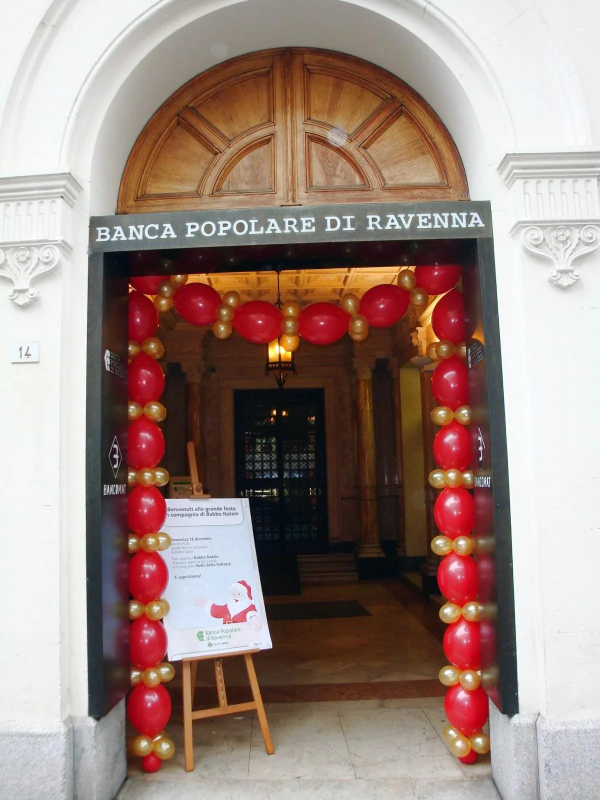 Ingresso della Banca Popolare di Ravenna incorniciato da colonne e da un arco di palloncini rossi e oro, con un cartello che annuncia la festa con Babbo Natale