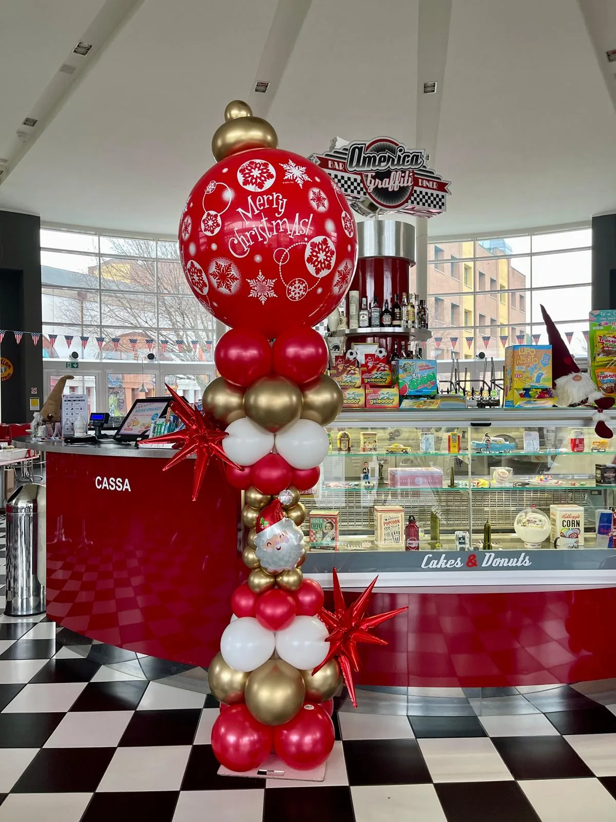 Colonna di palloncini rossi, oro e bianchi con stelle in mylar rosse, una faccina di Babbo Natale e in cima un grande palloncino rosso con la scritta Merry Christmas