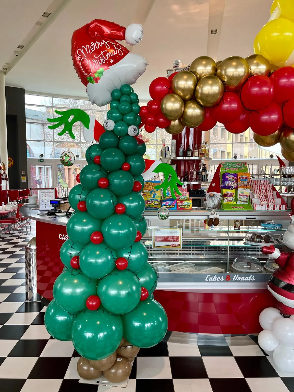 Albero di palloncini verdi con le palline rosse, gli occhi e le mani verdi, con in cima un cappello di Babbo Natale in mylar con la scritta Merry Christmas, accanto a una ghirlanda rossa e oro