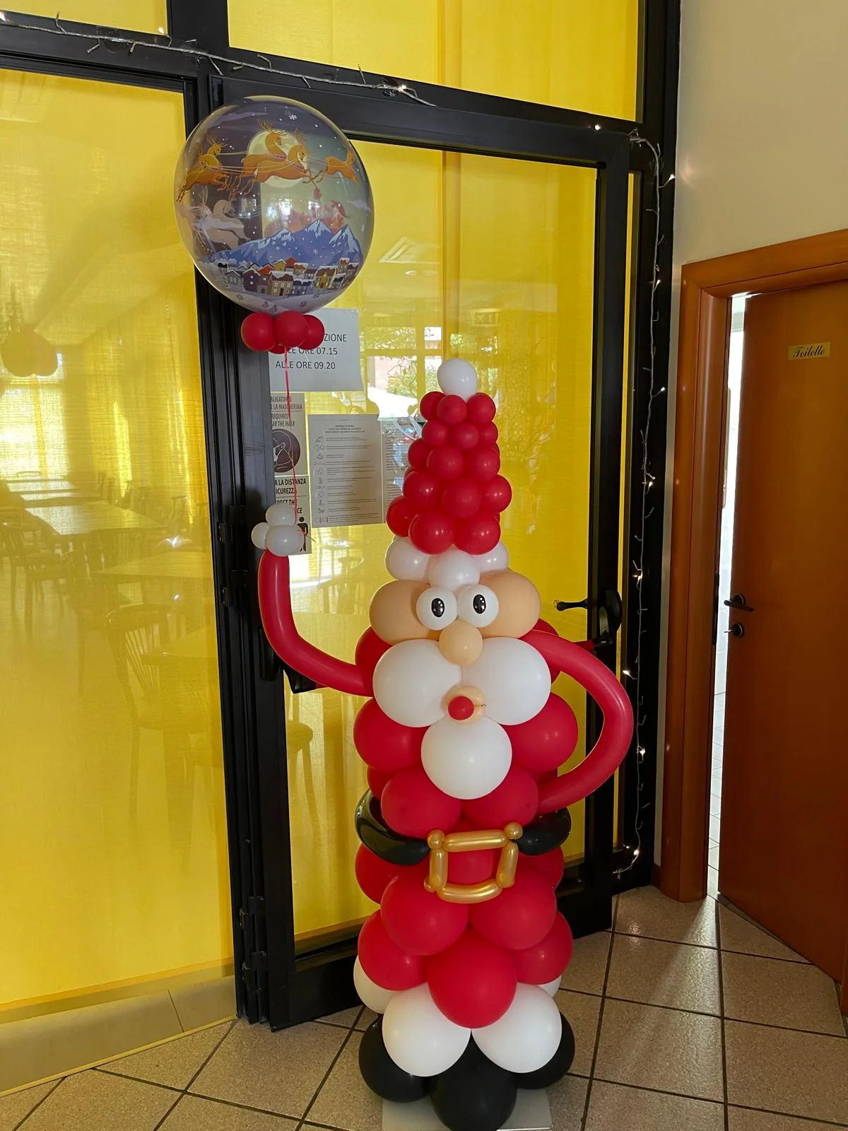 Babbo Natale di palloncini rossi e bianchi con la cintura dorata, che regge un palloncino trasparente con la slitta e le renne, davanti alla porta di una sala
