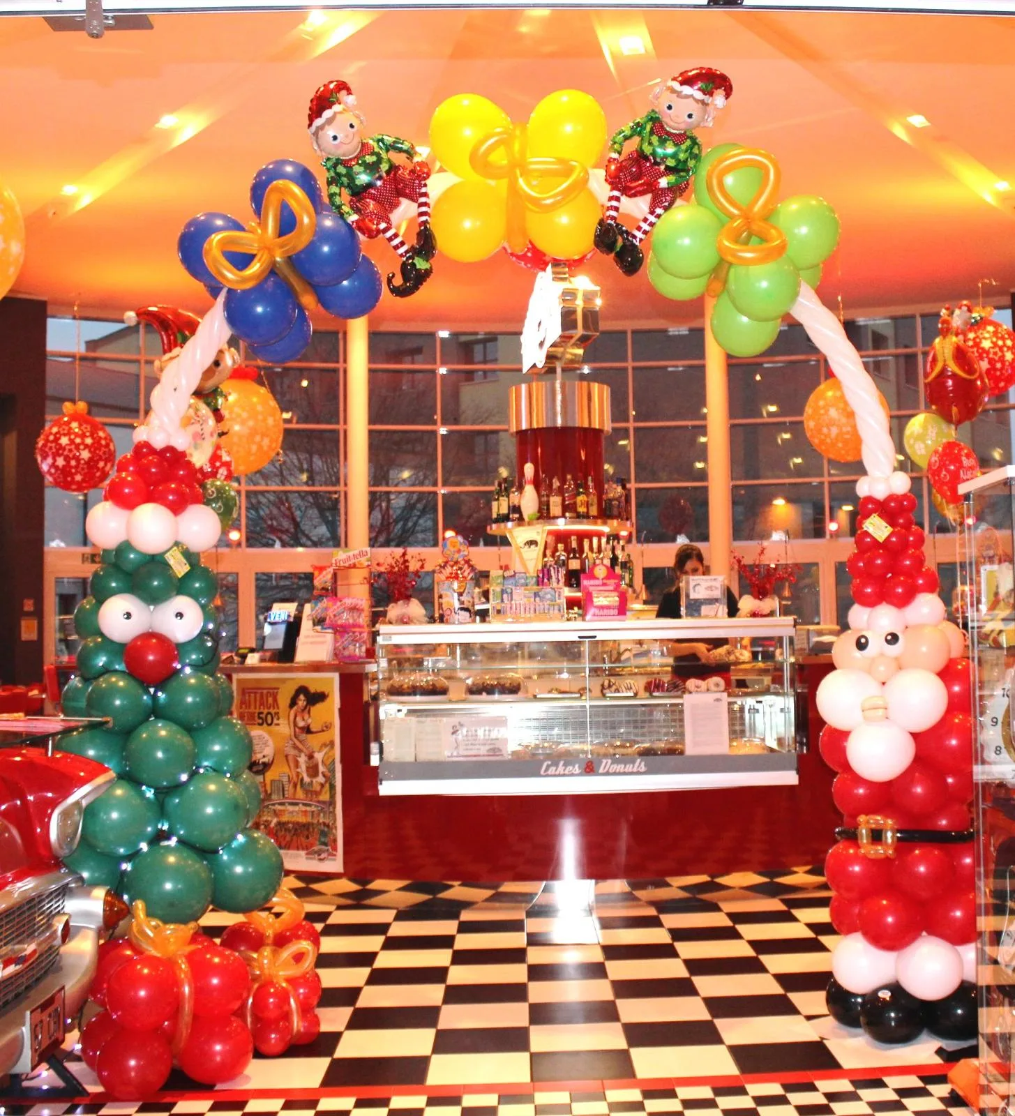 Arco di palloncini blu, gialli e verdi con due elfi in mylar, sorretto da un albero di Natale e da un Babbo Natale di palloncini, davanti al bancone di un locale