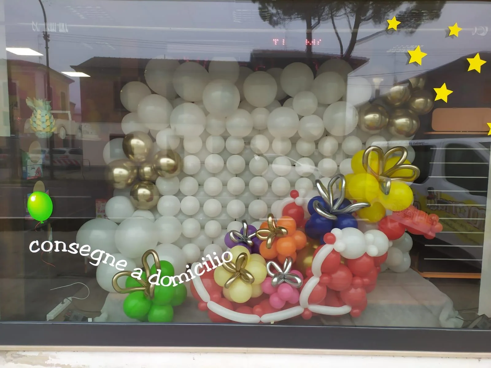Vetrina con una parete di palloncini bianchi e oro e pacchi regalo di palloncini colorati, con la scritta consegne a domicilio sul vetro