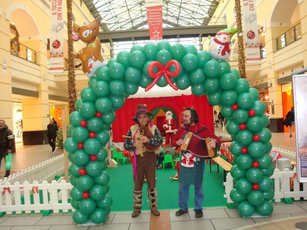 Arco di palloncini verdi con le palline rosse, un fiocco rosso, una renna e un pupazzo di neve in mylar, con due musicisti davanti, in una galleria commerciale