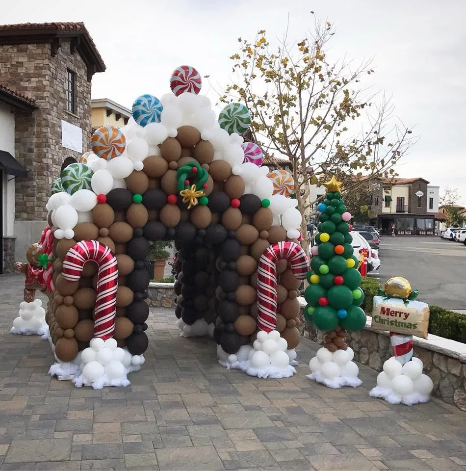 Casetta di pan di zenzero fatta di palloncini marroni e bianchi, con bastoncini di zucchero e caramelle giganti, accanto a un albero di palloncini e a un cartello Merry Christmas, all'aperto