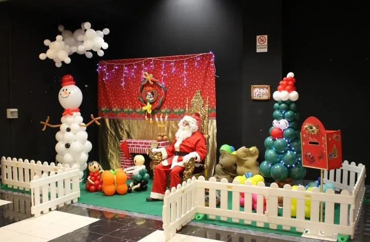 Villaggio di Natale in una galleria: Babbo Natale sul trono, pupazzo di neve e albero di palloncini, cassetta delle lettere e staccionata bianca