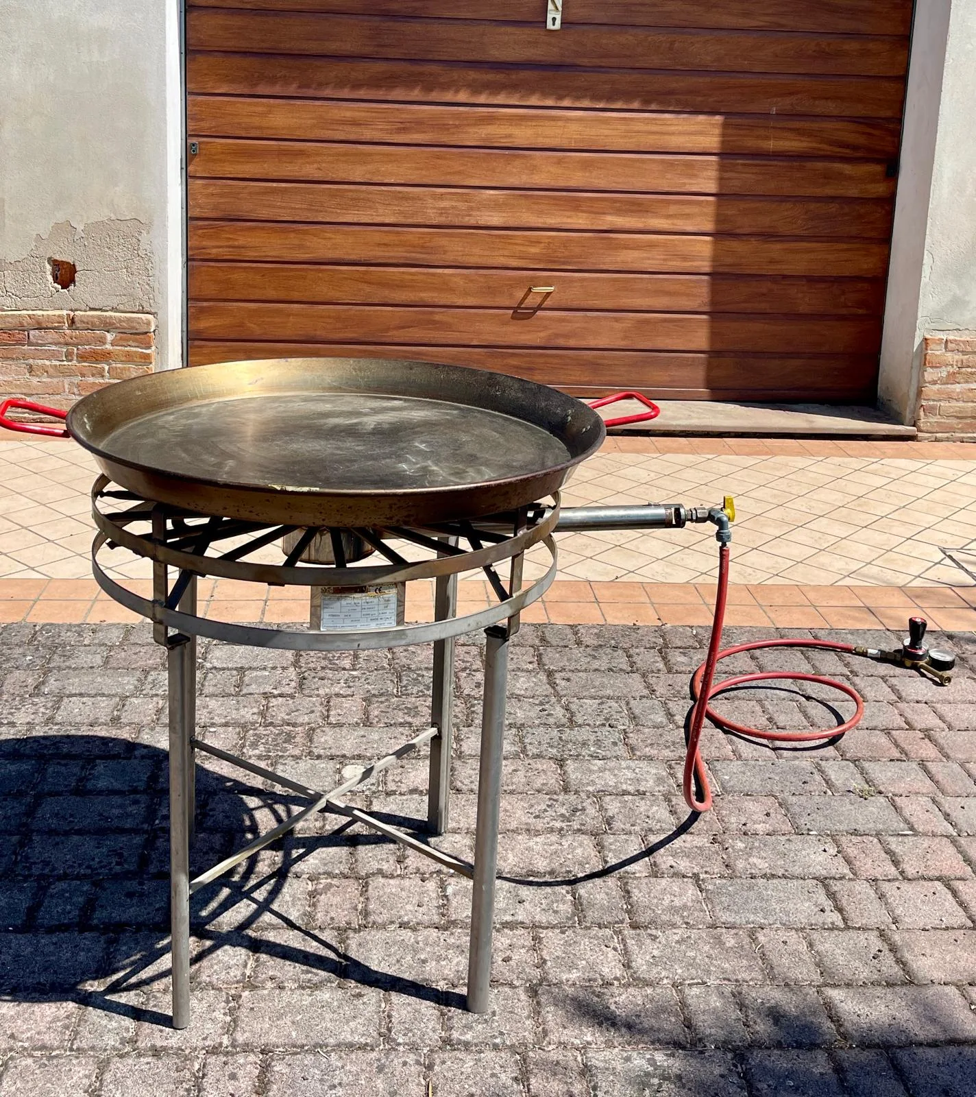 Grande padella da paella in acciaio con manici rossi appoggiata sul fornellone a gas con treppiede e tubo di collegamento alla bombola