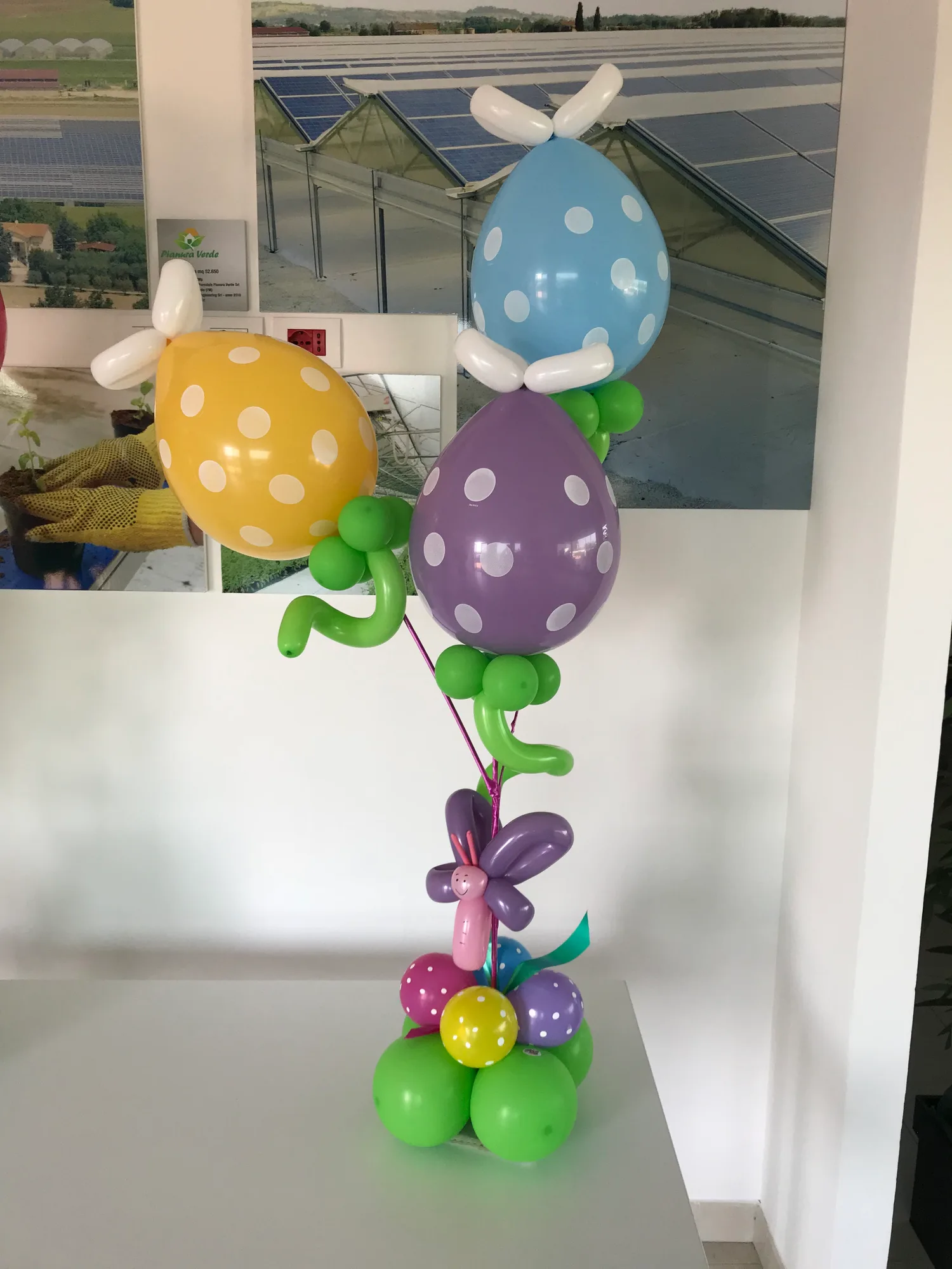 Composizione da tavolo con tre palloncini a forma di uovo a pois gialli, azzurri e viola, foglie verdi e una farfalla di palloncini