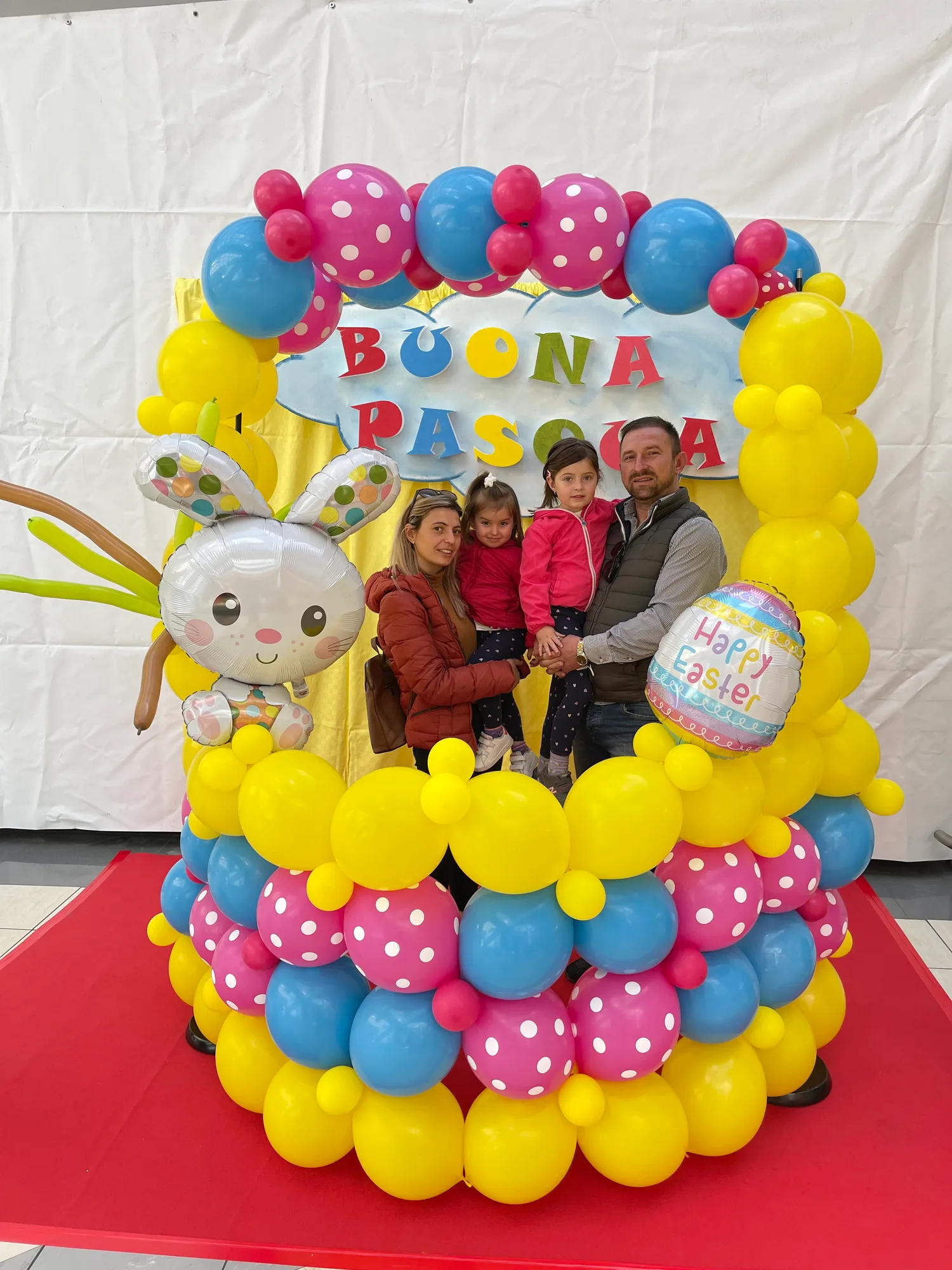 Cornice di palloncini gialli, azzurri e fucsia a pois con un coniglietto e un uovo Happy Easter in mylar e la scritta Buona Pasqua, con una famiglia in posa dentro