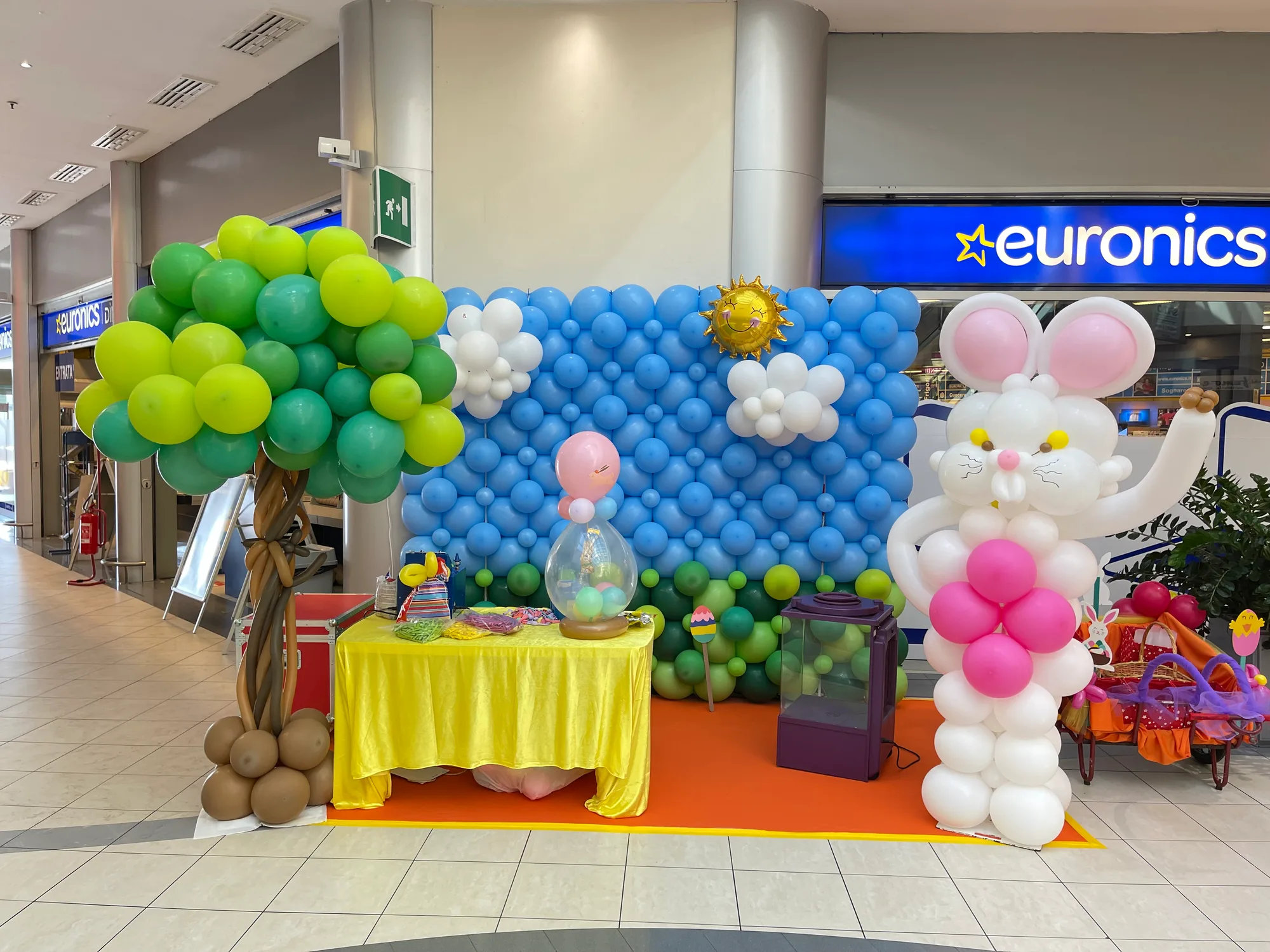 Parete di palloncini azzurri e verdi con nuvole e sole, un albero e un coniglio bianco e rosa di palloncini, davanti a un negozio in una galleria commerciale