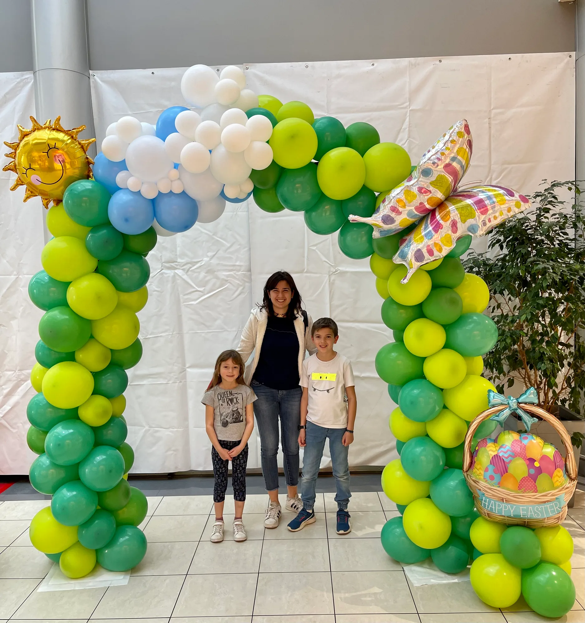 Arco di palloncini verdi, azzurri e bianchi con un sole, una farfalla e un cestino di uova Happy Easter in mylar, con una ragazza e due bambini sotto