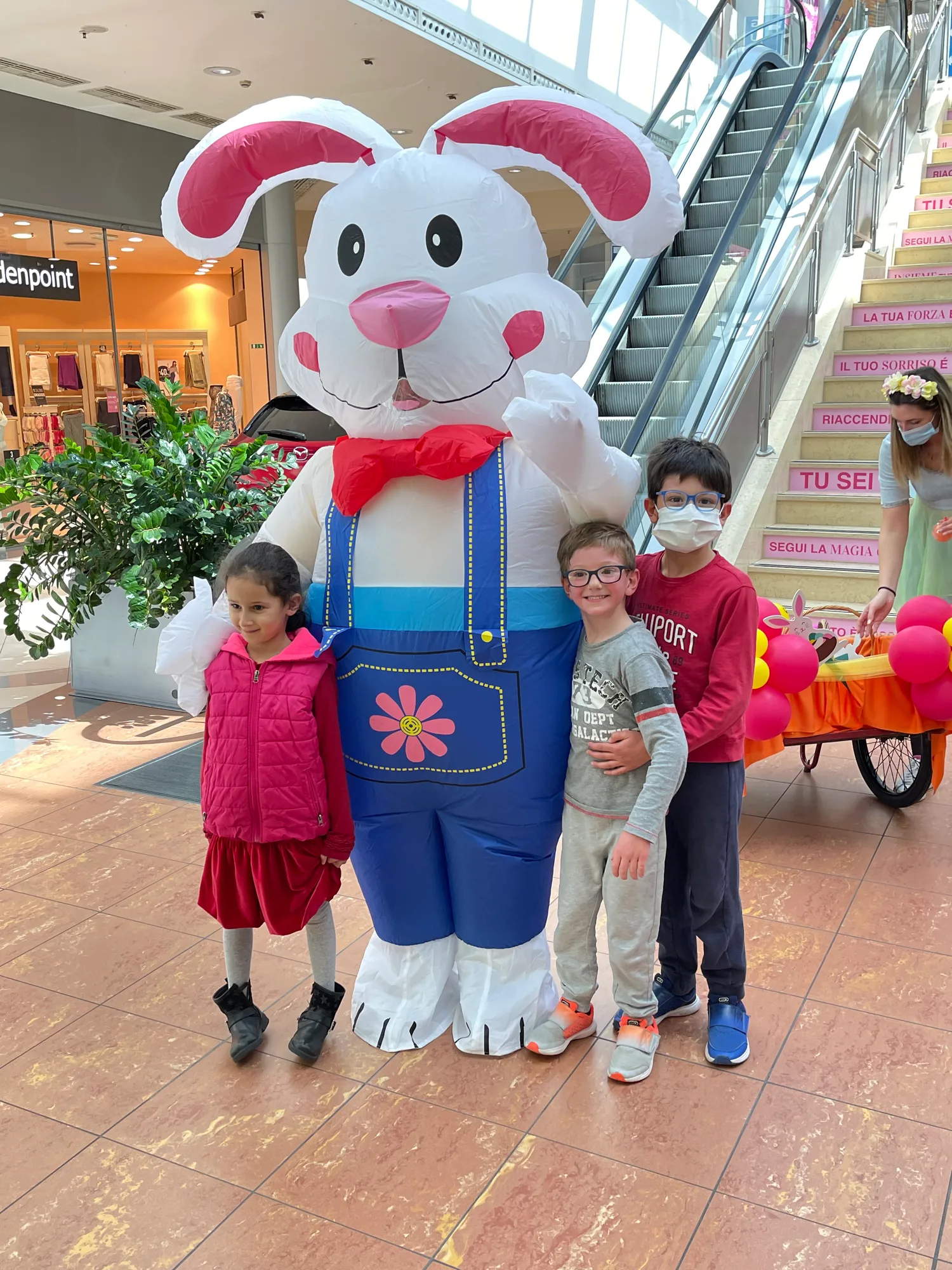 Tre bambini in posa accanto alla mascotte gonfiabile di un coniglio bianco con la salopette blu, in una galleria commerciale