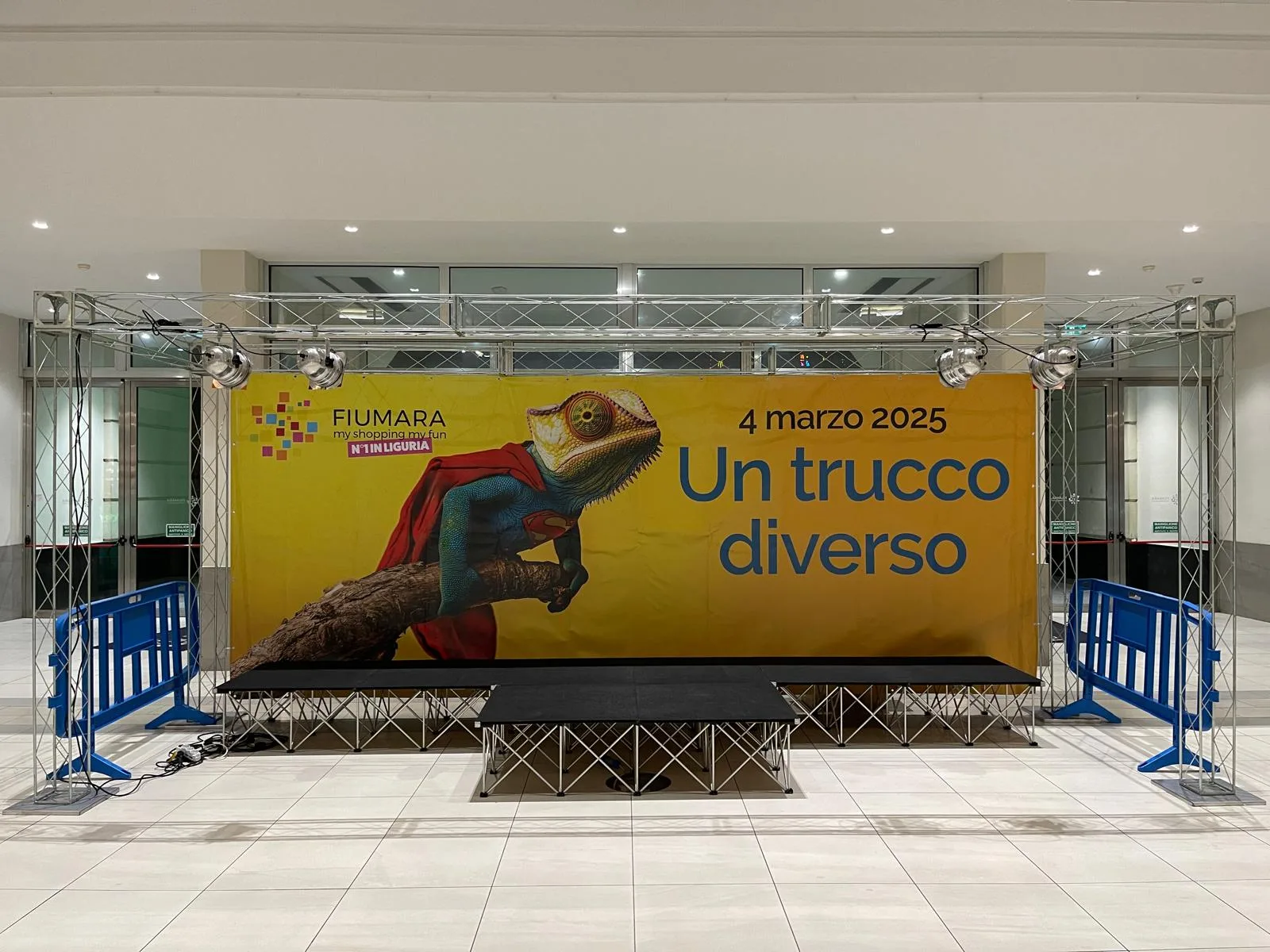 Palco modulare nero alto 40 cm montato in una galleria commerciale davanti a un fondale giallo stampato, con traliccio, fari e transenne blu ai lati