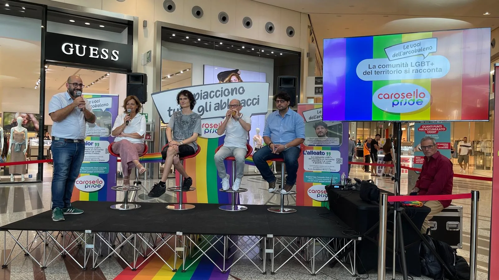 Palco modulare nero con schermo televisivo acceso, casse e sgabelli, allestito per una conferenza in una galleria commerciale