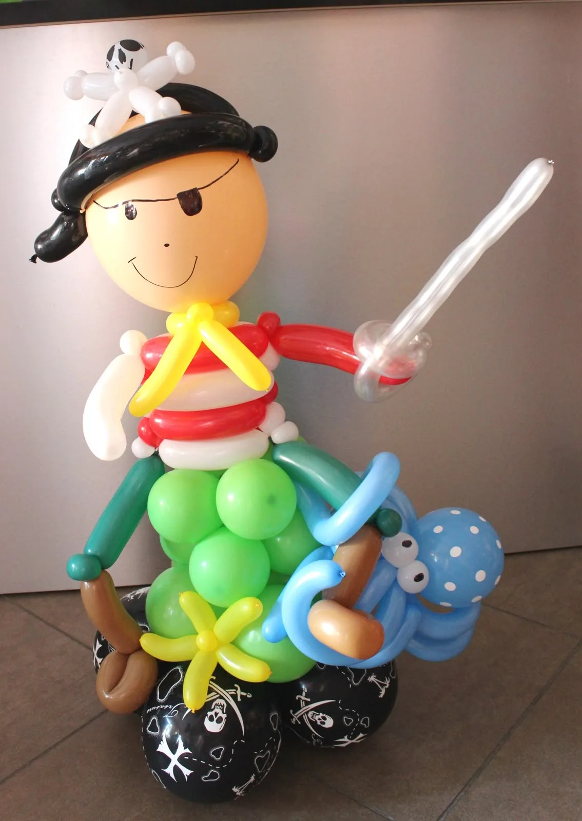 Pirata di palloncini con la benda sull'occhio, la spada, la maglia a righe rosse e bianche e un polpo azzurro a pois, su una base di palloncini neri con i teschi