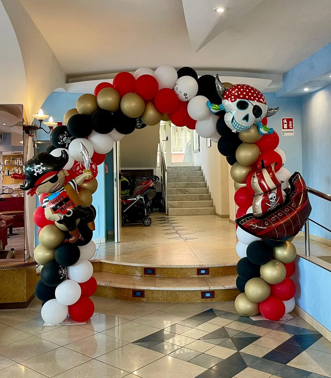 Arco di palloncini rossi, neri, bianchi e oro con un pirata, un teschio con la bandana e un veliero in mylar, all'ingresso di una sala