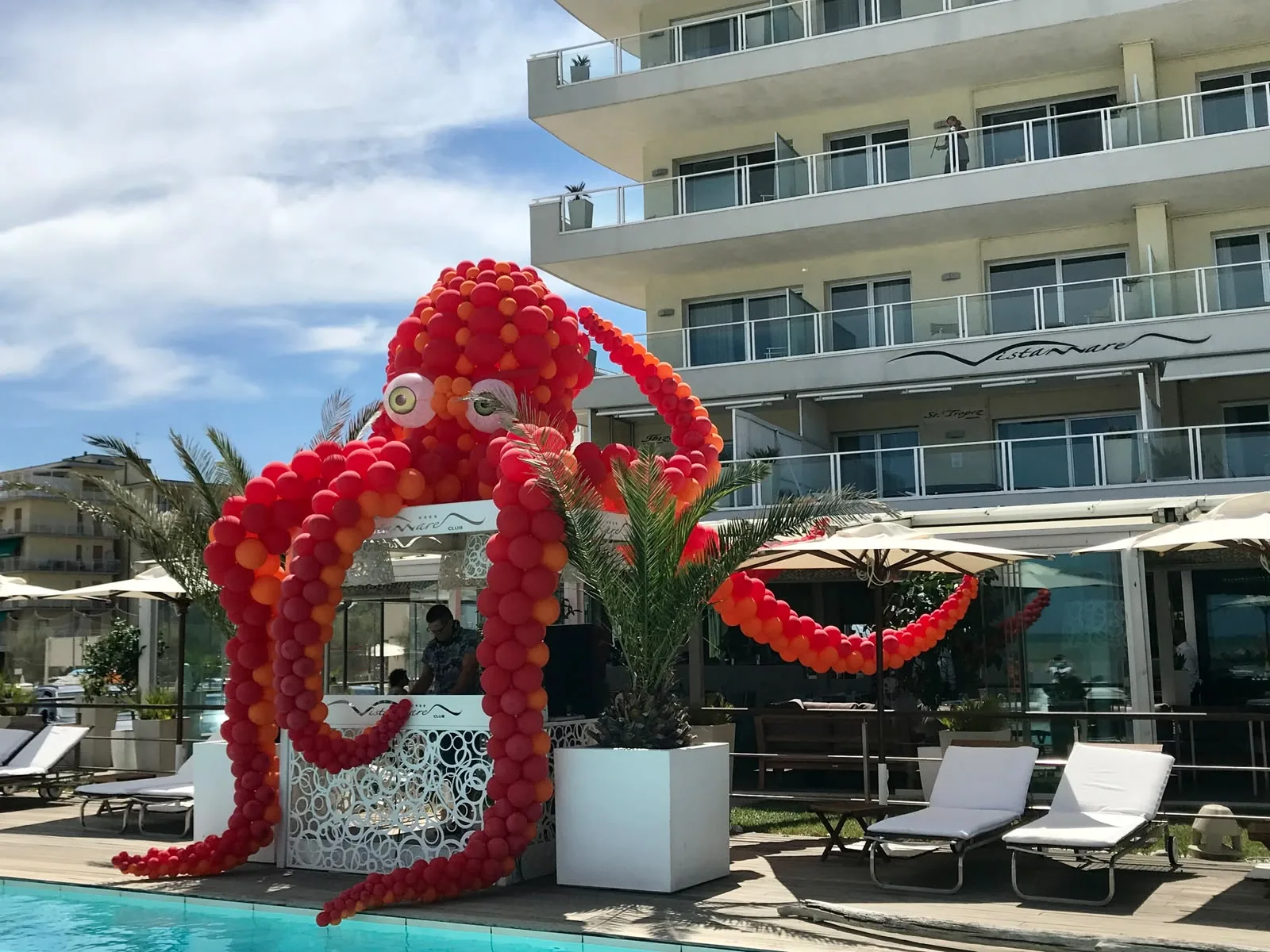 Polpo gigantesco di palloncini rossi e arancioni con i tentacoli che avvolgono una postazione a bordo piscina, davanti a un hotel