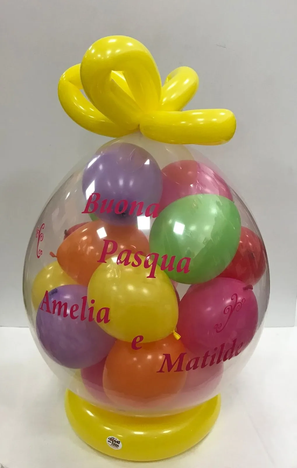 Grande palloncino trasparente pieno di palloncini colorati con la scritta stampata Buona Pasqua Amelia e Matilde