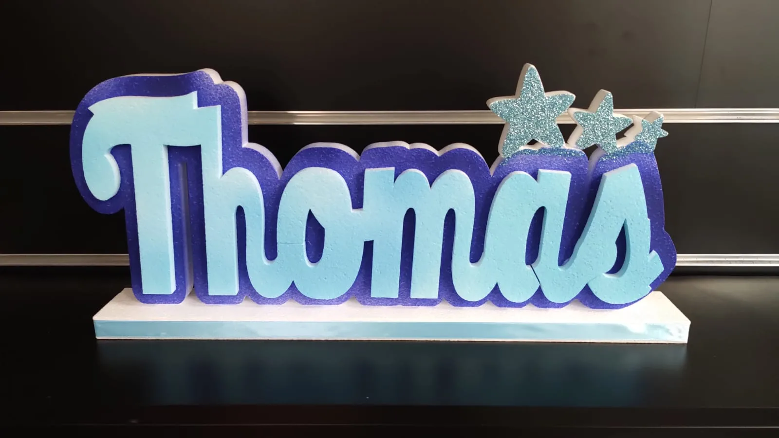 Nome Thomas in lettere di polistirolo azzurre e blu con tre stelle glitterate, su una base bianca e azzurra
