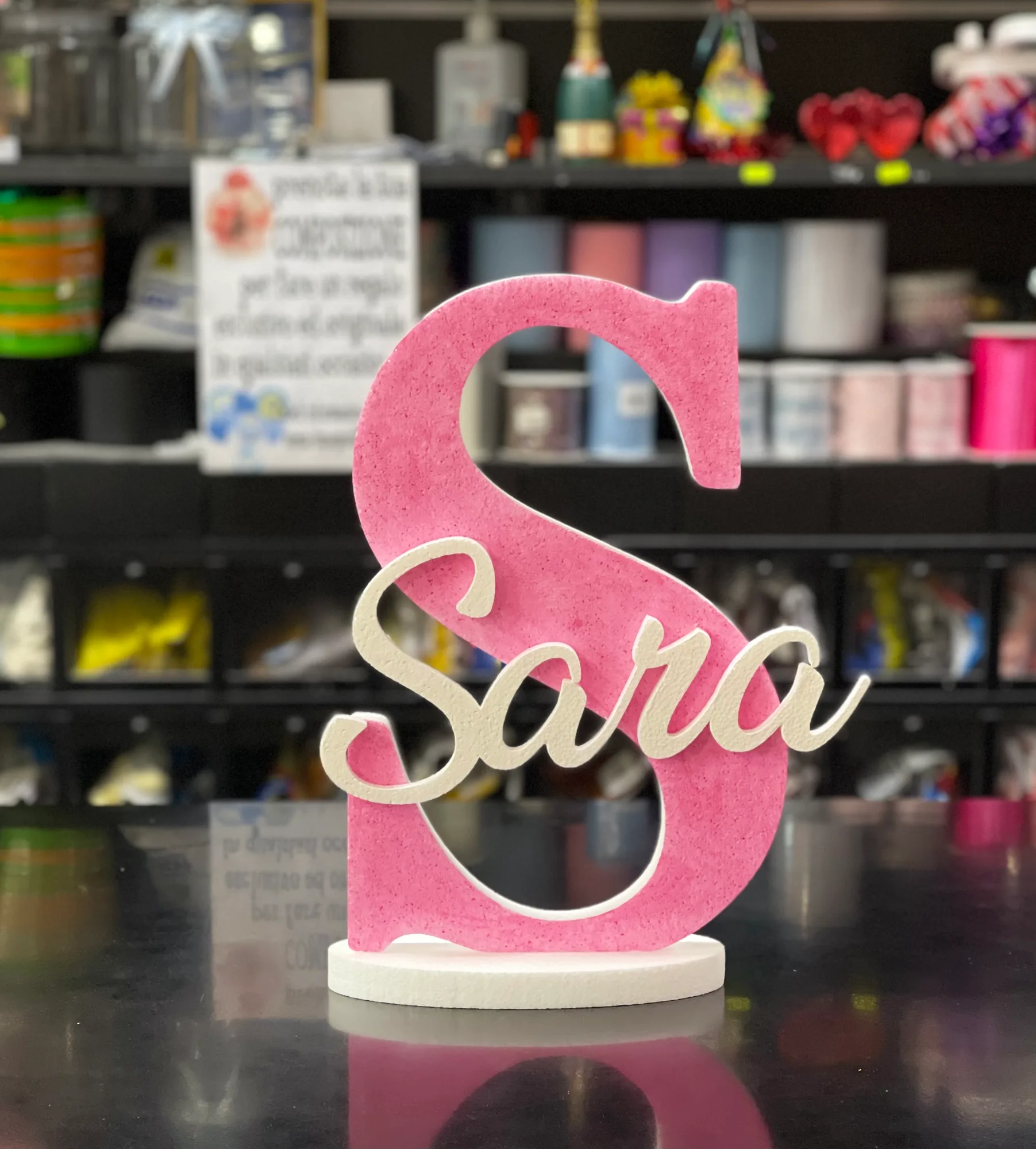 Grande lettera S in polistirolo rosa con il nome Sara in corsivo bianco, su una base tonda bianca