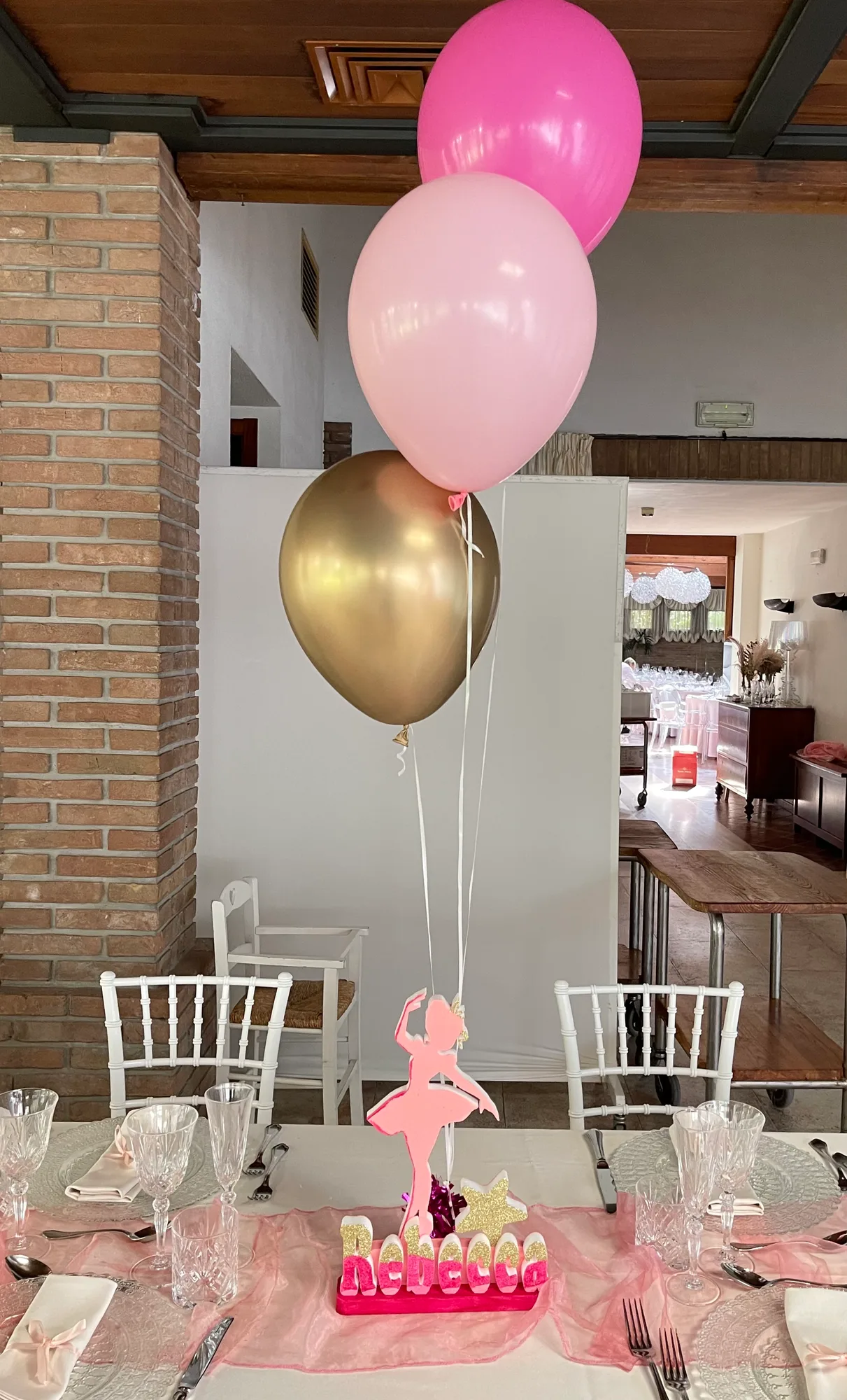 Centrotavola con una ballerina in polistirolo rosa, il nome Rebecca glitterato e tre palloncini rosa e oro, su una tavola apparecchiata con runner rosa