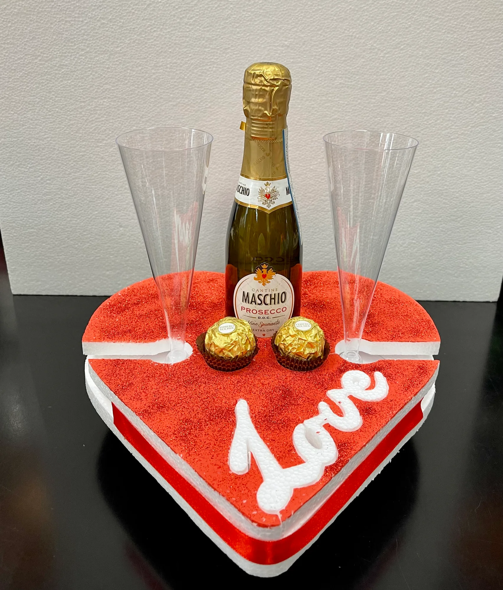 Vassoio a forma di cuore in polistirolo rosso glitterato con la scritta Love, che regge una bottiglia di spumante, due flûte e due cioccolatini
