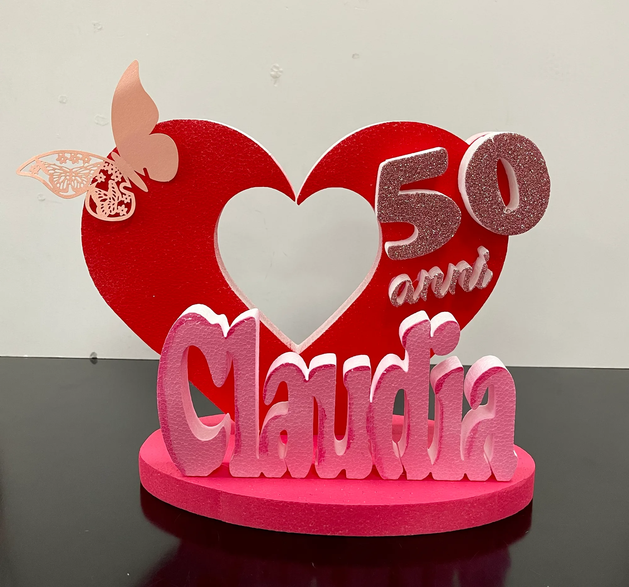 Grande cuore rosso in polistirolo con un cuore vuoto al centro, una farfalla rosa, la scritta 50 anni glitterata e il nome Claudia in lettere rosa su una base fucsia