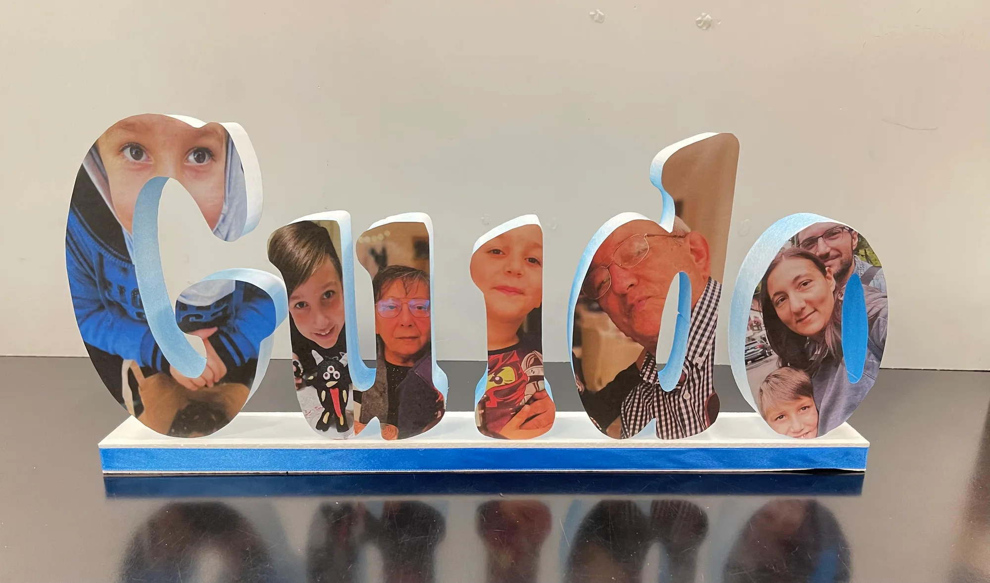 Nome Guido in lettere di polistirolo ricoperte di fotografie di familiari, su una base bianca con nastro azzurro