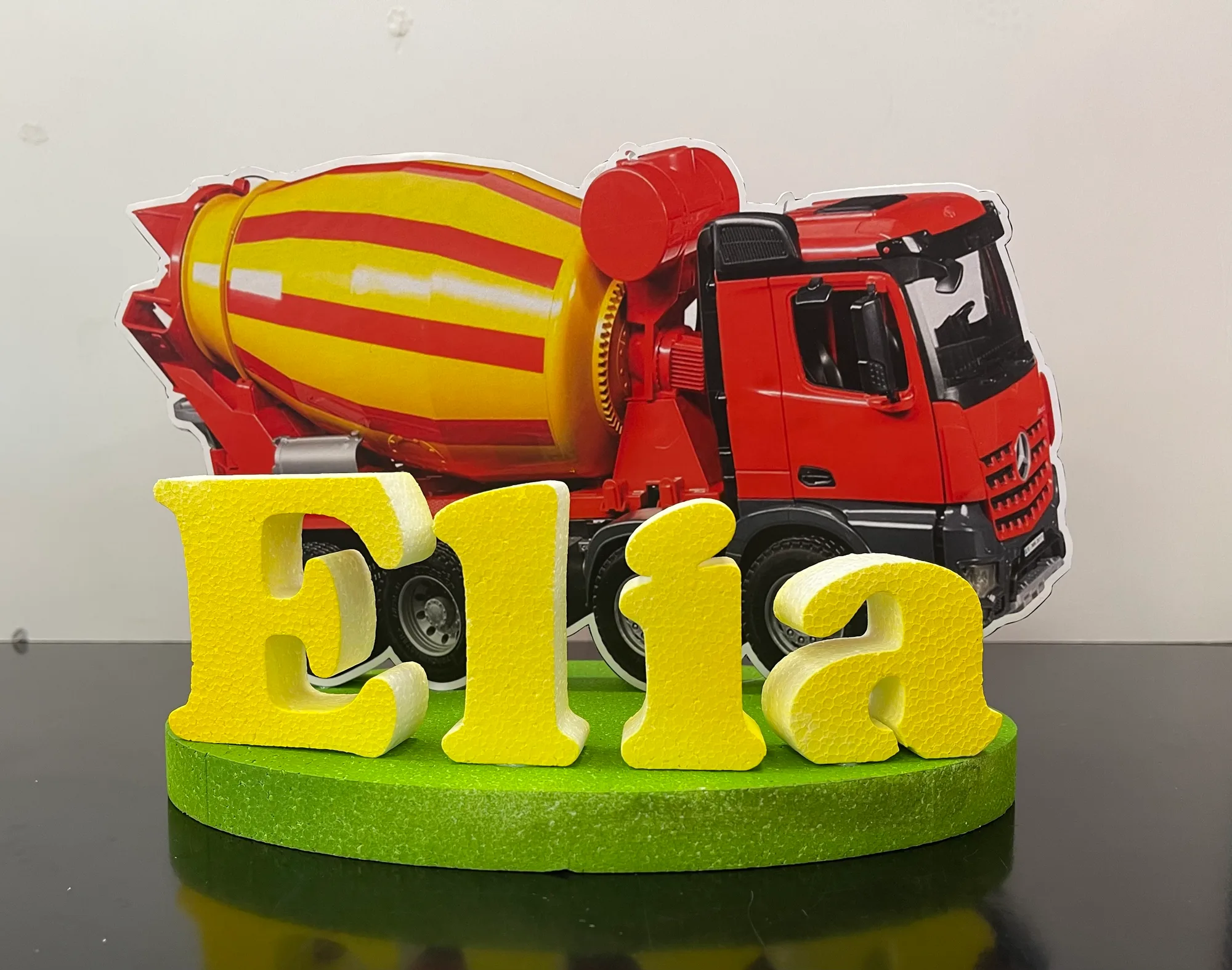 Sagoma stampata di una betoniera rossa e gialla con il nome Elia in lettere di polistirolo gialle su una base verde