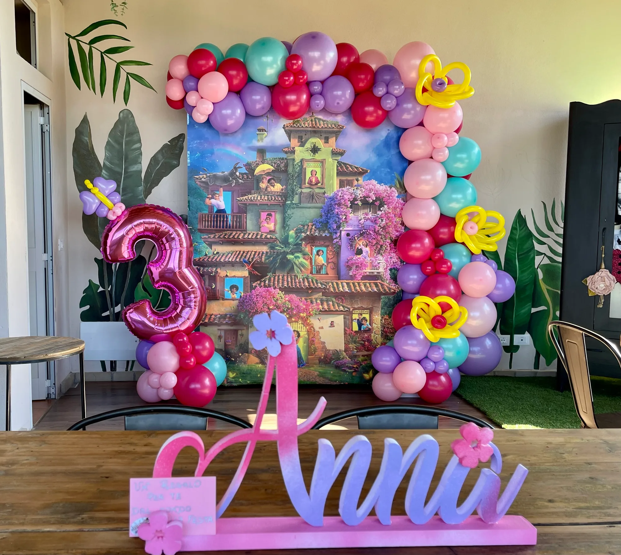 Nome Anna in polistirolo rosa e lilla con fiori, sul tavolo davanti a un fondale stampato incorniciato da palloncini rosa, viola e verde acqua e da un numero 3 fucsia