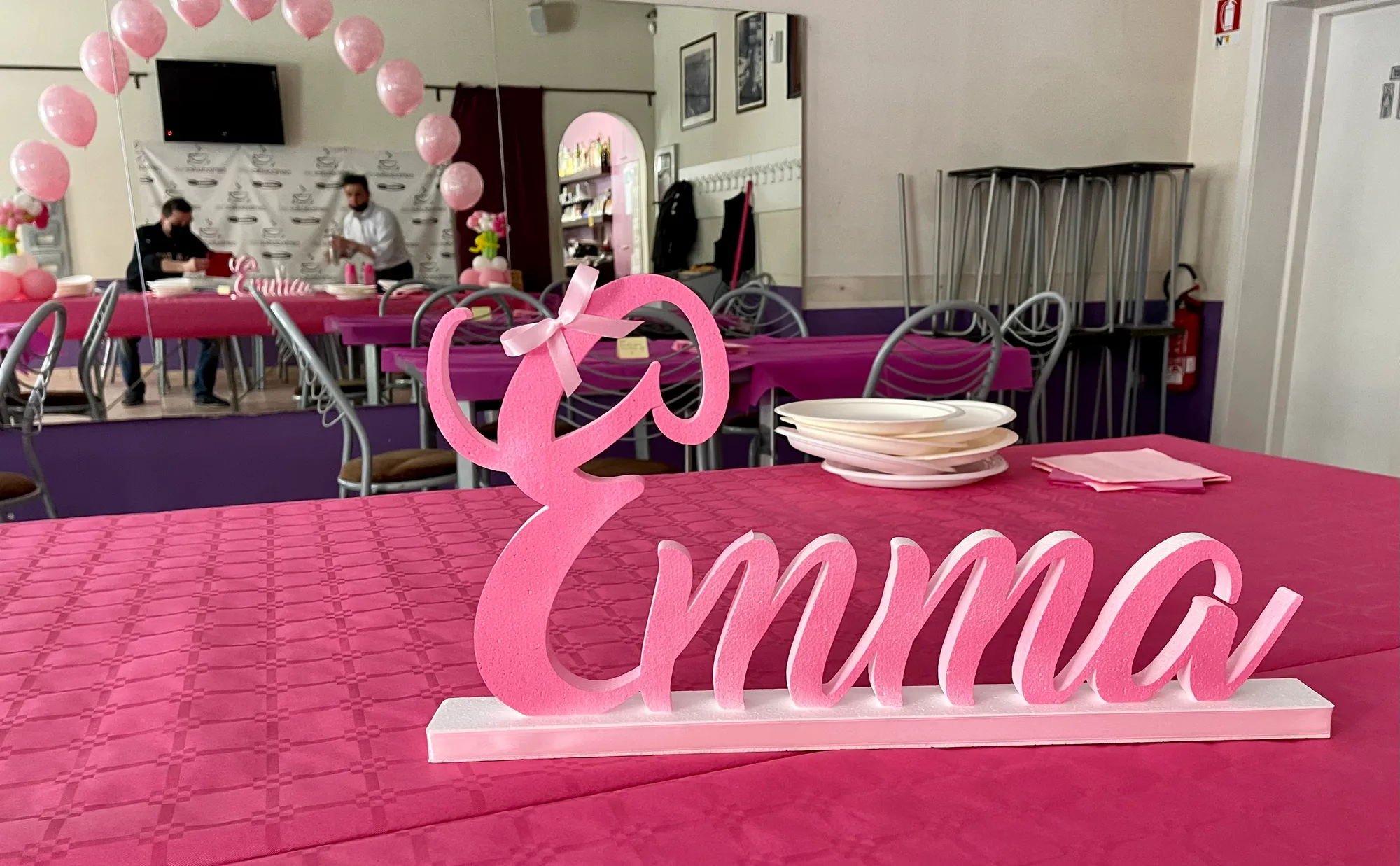 Nome Emma in polistirolo rosa in corsivo con un fiocco rosa, su un tavolo apparecchiato con tovaglia fucsia