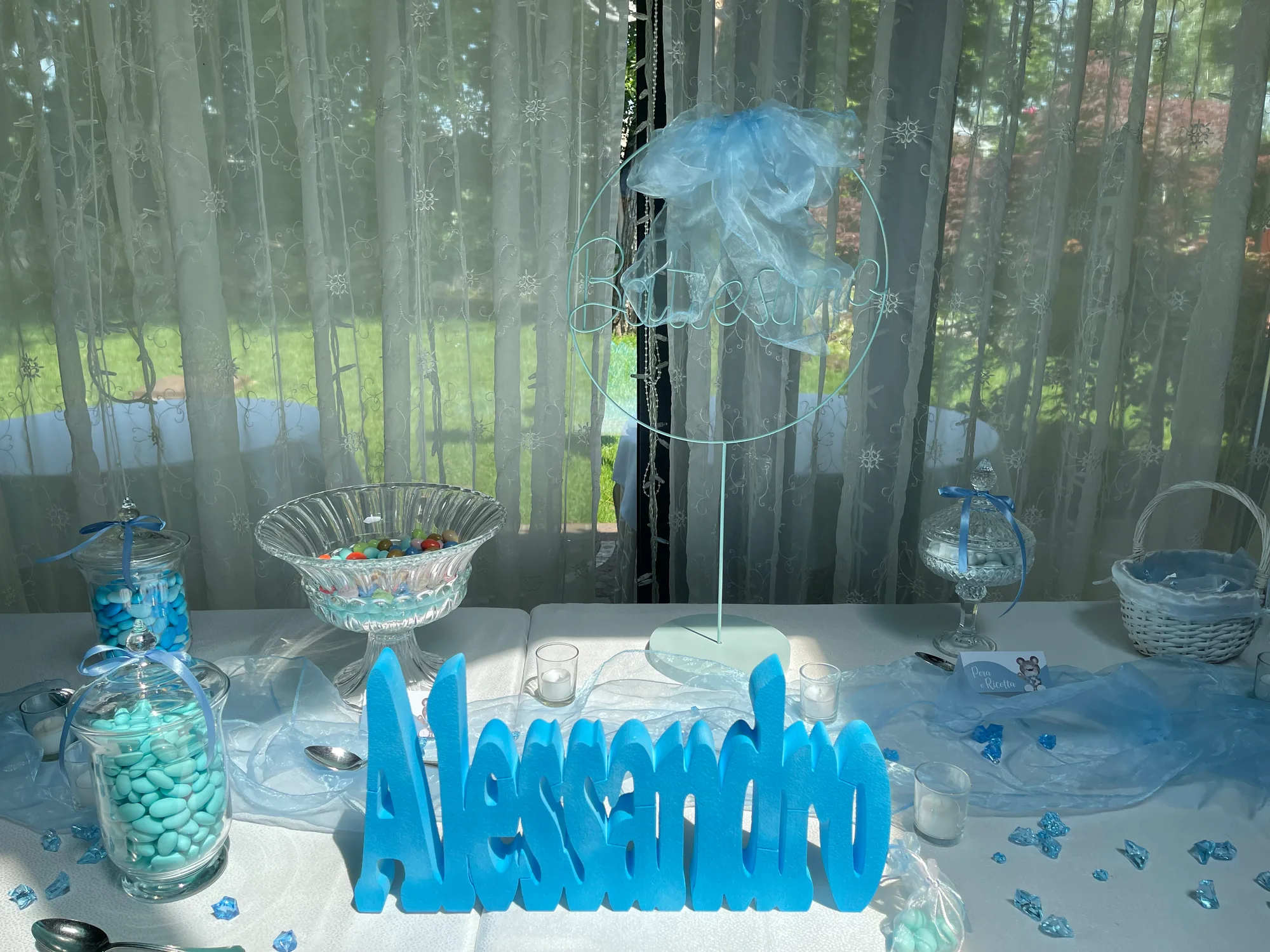 Nome Alessandro in lettere di polistirolo azzurre sul tavolo dei confetti di un battesimo, con barattoli di confetti azzurri e candele