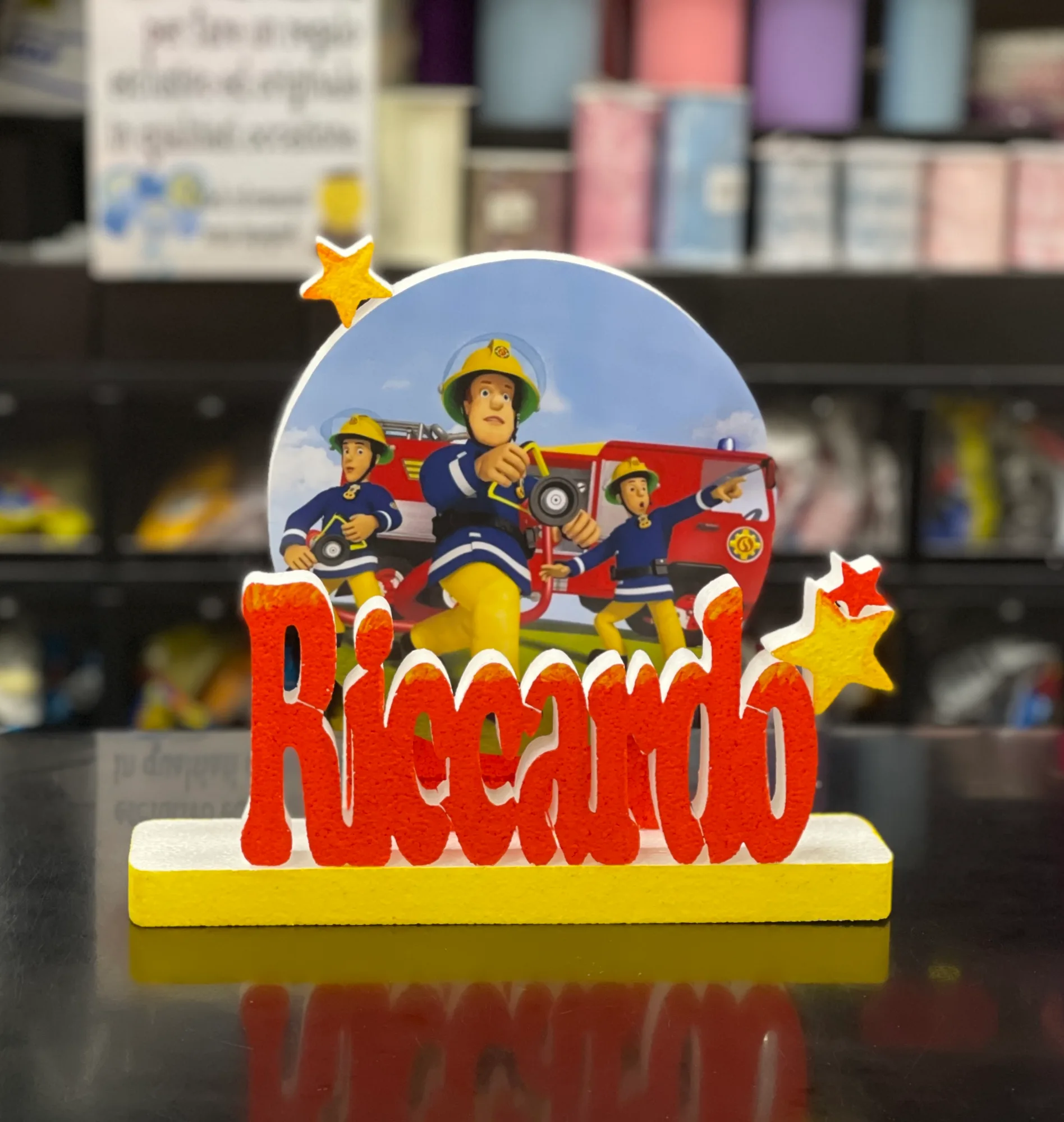 Sagoma tonda stampata con tre pompieri dei cartoni e il camion rosso, con il nome Riccardo in lettere rosse e stelle gialle su una base gialla