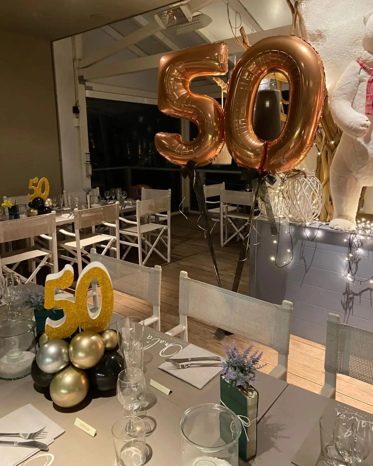 Centrotavola con il numero 50 in polistirolo oro glitterato su palloncini oro, argento e neri, e due grandi palloncini 50 in mylar color rame in una sala apparecchiata