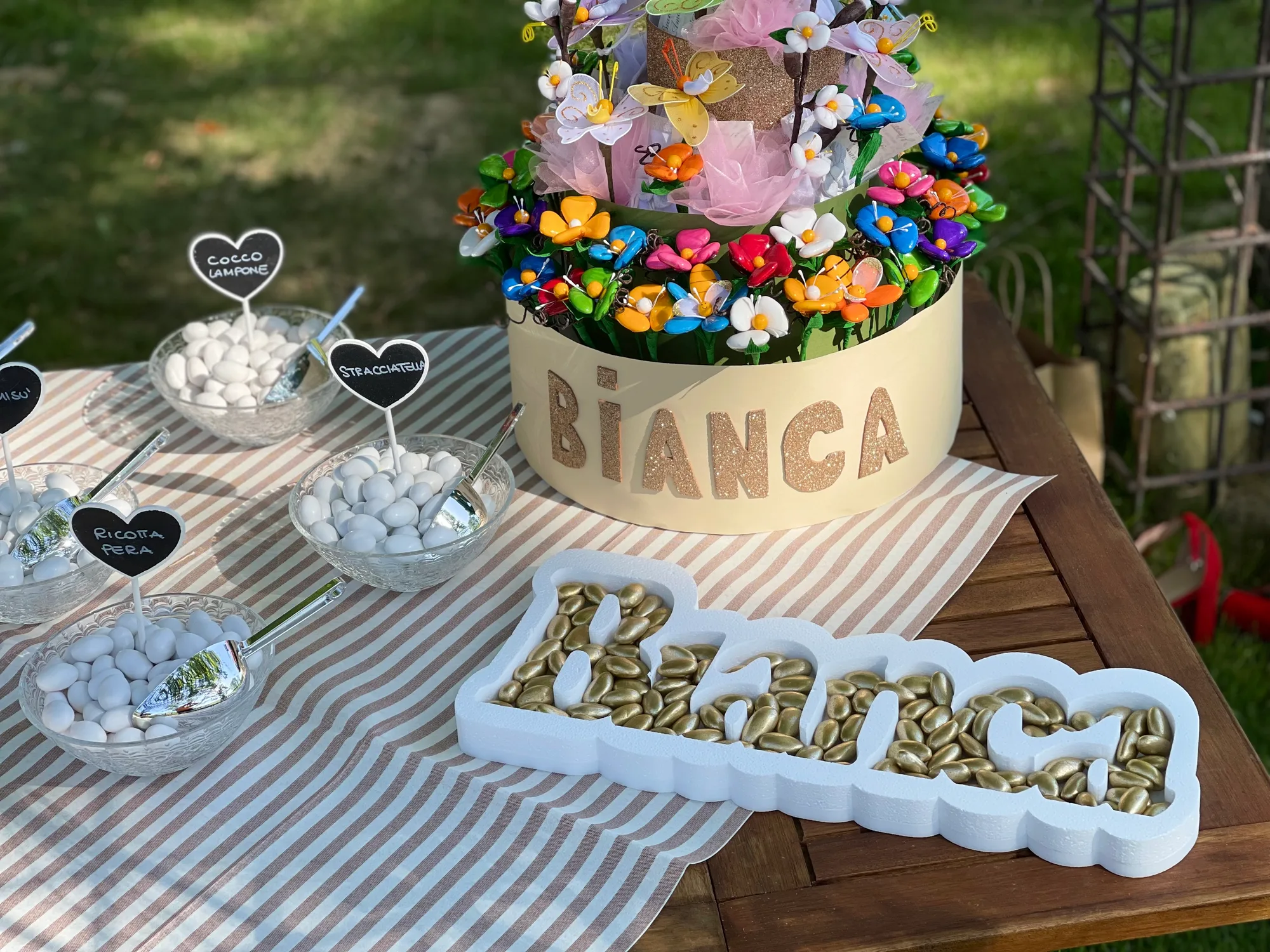 Portaconfetti in polistirolo bianco con il nome Bianca riempito di confetti dorati, su un tavolo con ciotole di confetti e una torta di fiori con la scritta BIANCA glitterata