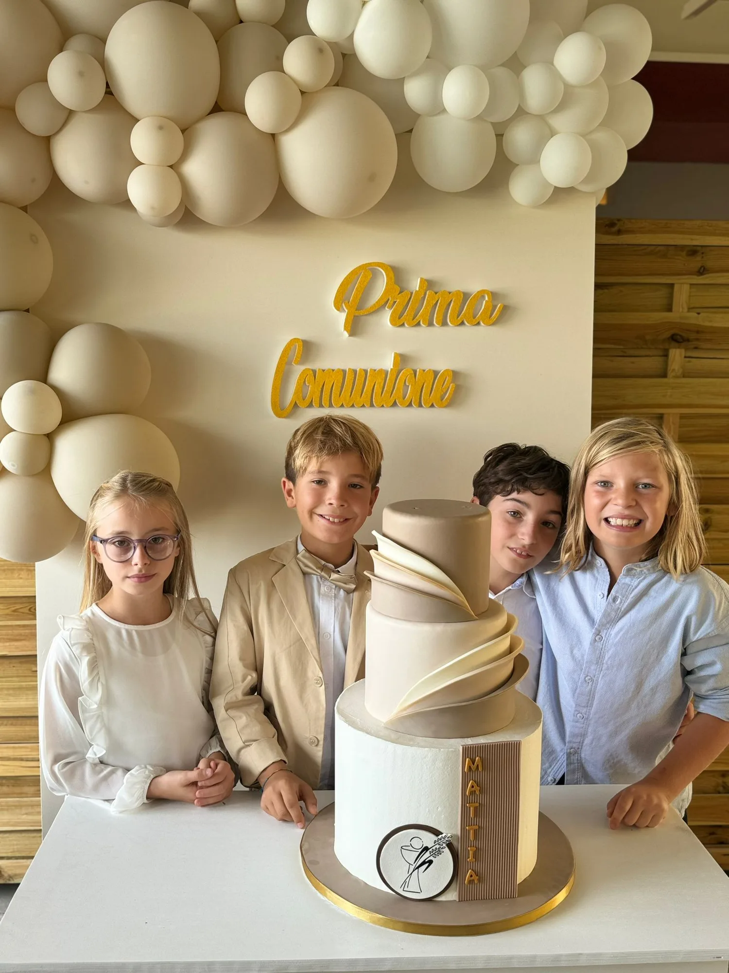 Scritta Prima Comunione in corsivo oro glitterato su un pannello con palloncini panna, dietro a quattro bambini in posa accanto alla torta
