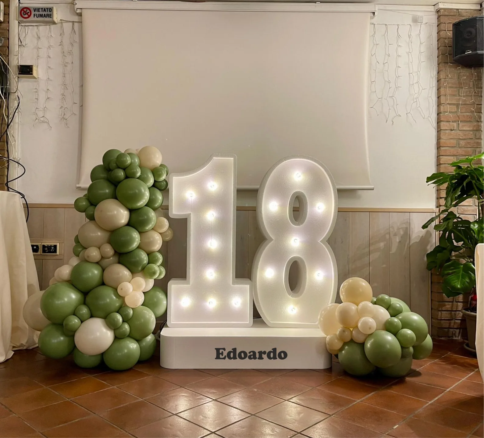 Numero 18 luminoso a lampadine su base con il nome Edoardo, affiancato da ghirlande di palloncini verde salvia e panna in una sala con pavimento in cotto