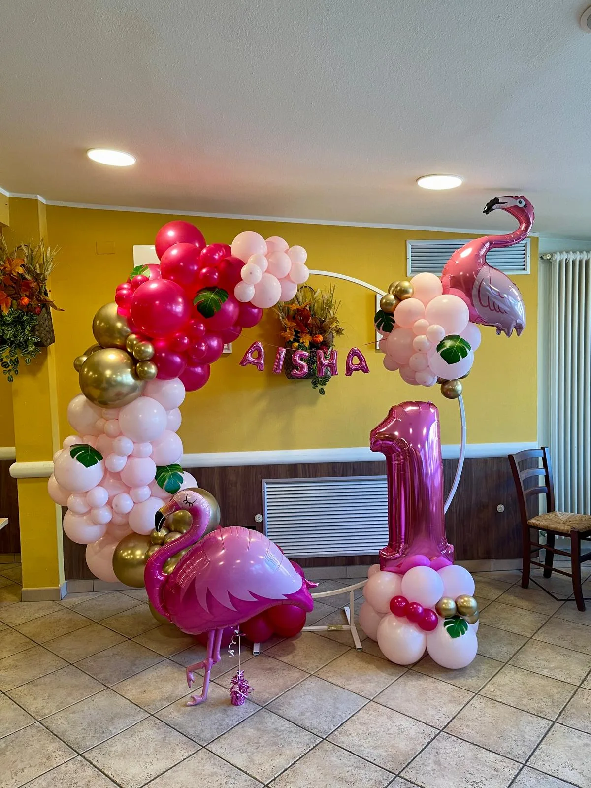Cerchio di palloncini fucsia, rosa e oro con fenicotteri, foglie tropicali, scritta Aisha e numero 1 fucsia, allestito in sala davanti a una parete gialla