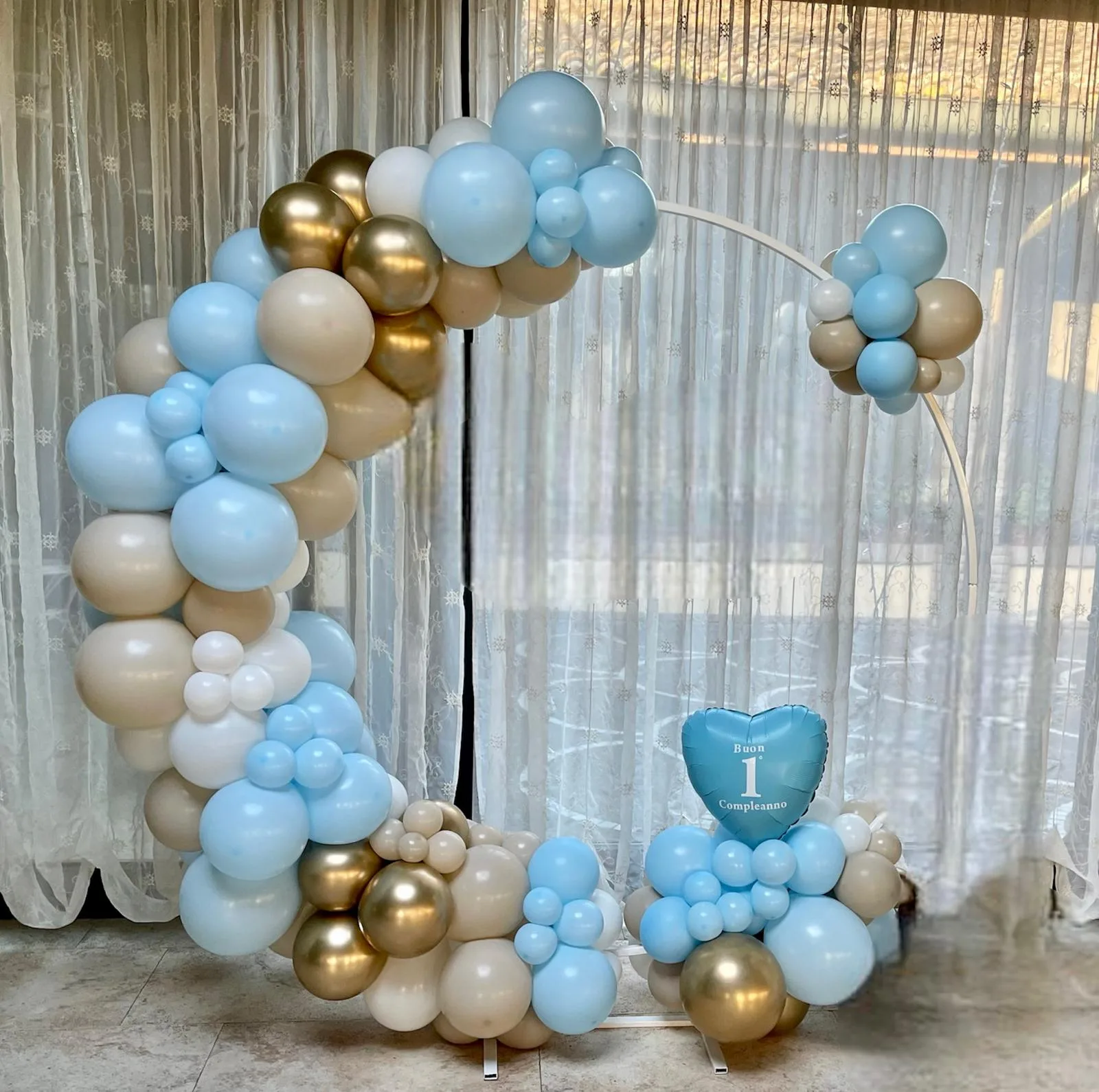 Cerchio di palloncini azzurri, sabbia, bianchi e oro con cuore azzurro Buon 1° Compleanno, davanti a una vetrata con tende