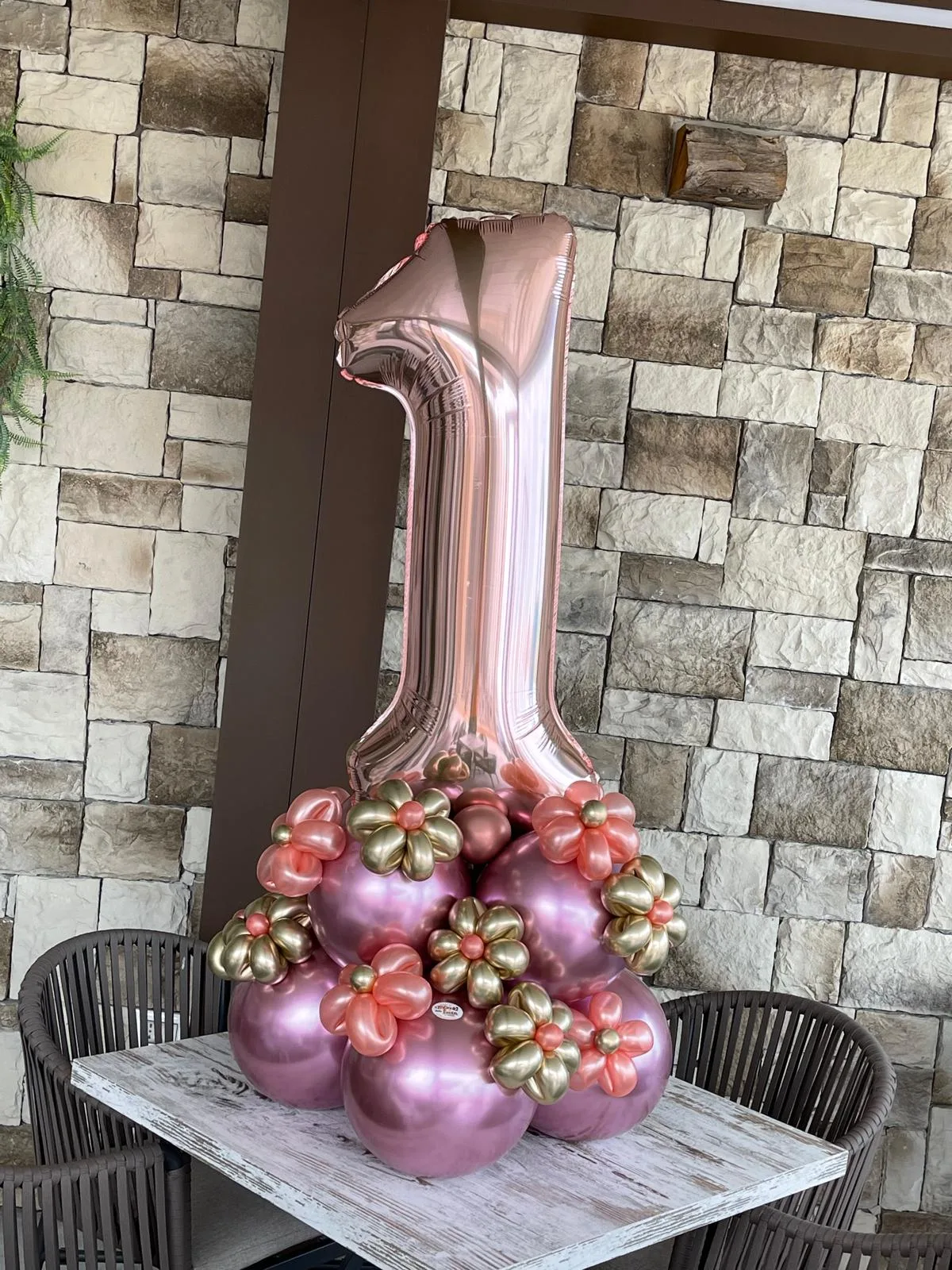 Numero 1 rosa gold in mylar su una base di palloncini rosa cromati con fiorellini oro e pesca, su un tavolino davanti a un muro in pietra