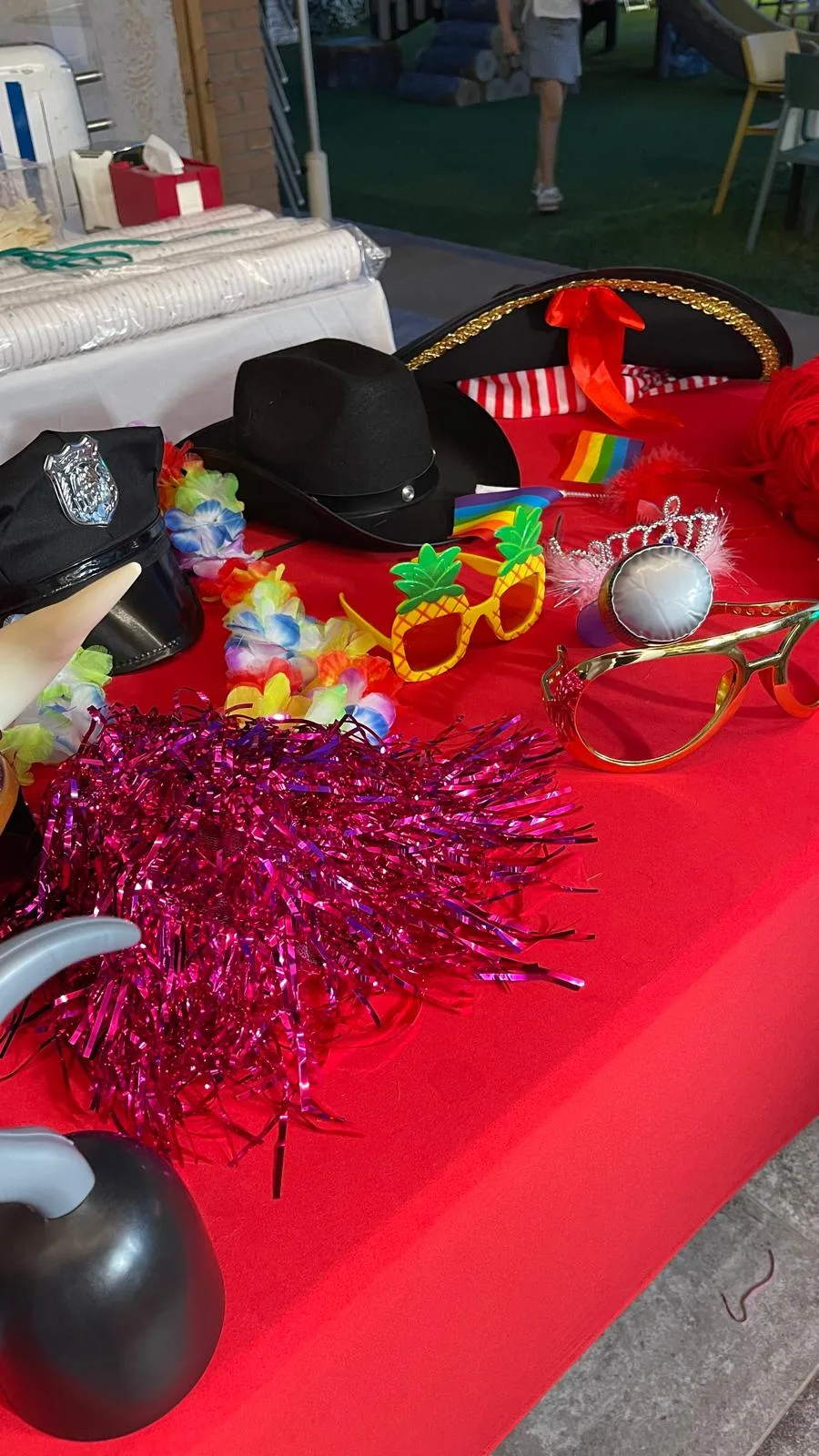 Tavolo rosso con gli accessori del photobooth: cappelli da cowboy e da poliziotto, cappello da pirata, occhiali a forma di ananas, corone, collane hawaiane e boa colorati