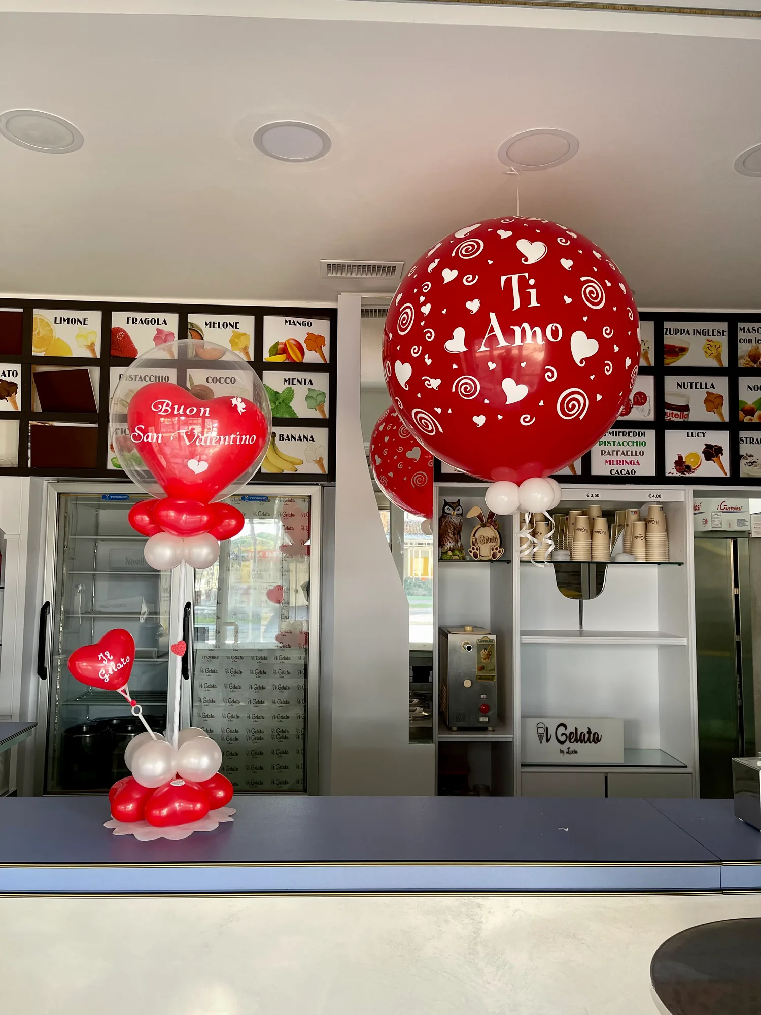 Composizione con un cuore rosso Buon San Valentino dentro un palloncino trasparente e un grande palloncino rosso con la scritta Ti Amo appeso sopra il bancone di una gelateria