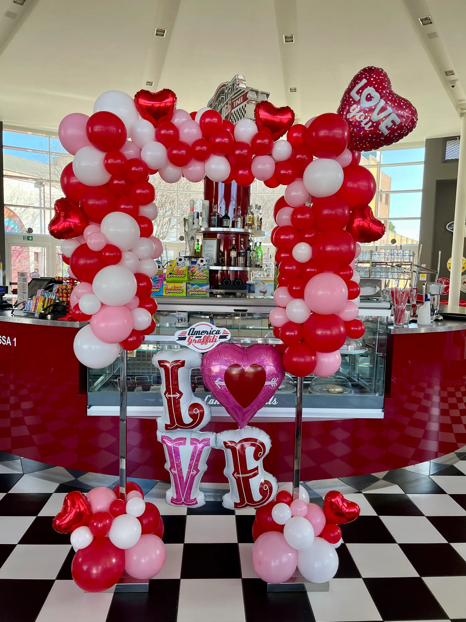 Cornice di palloncini rossi, rosa e bianchi con cuori in mylar, il cuore Love you e la scritta LOVE in lettere di mylar, davanti al bancone di un locale