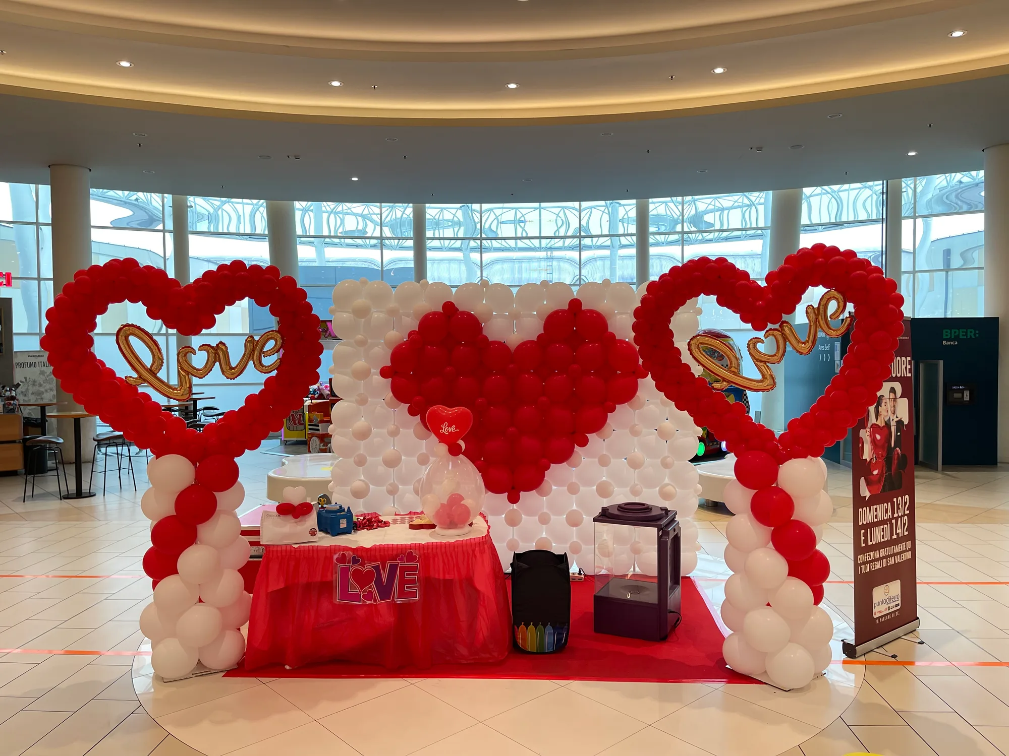 Allestimento di San Valentino in una galleria: due grandi cuori di palloncini rossi con la scritta love in mylar oro sulle colonne bianche e rosse, e al centro una parete di palloncini bianchi con un cuore rosso