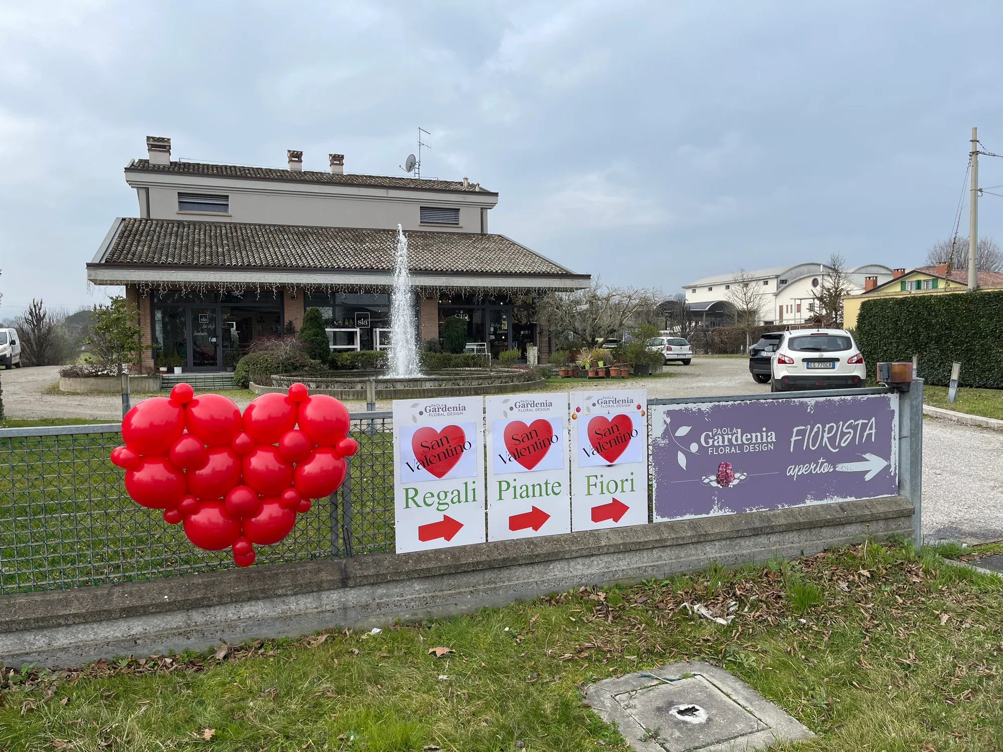 Cuore di palloncini rossi appeso alla recinzione di un vivaio, accanto ai cartelli San Valentino regali, piante e fiori