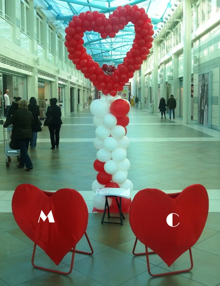 Grande cuore di palloncini rossi su una colonna di palloncini bianchi e rossi in una galleria, con davanti due sedie a forma di cuore con le lettere M e C