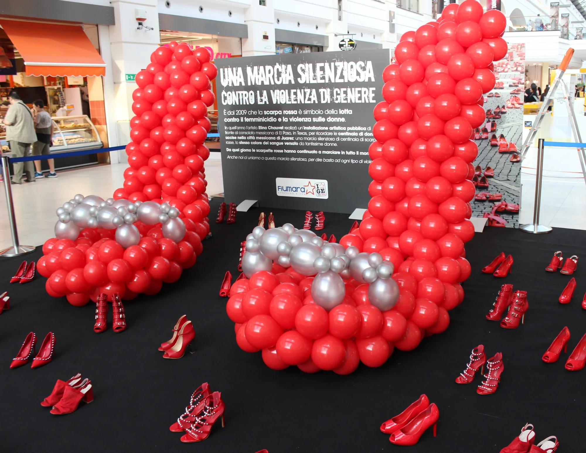 Due scarpe col tacco alte oltre due metri costruite con palloncini rossi e argento, allestite in un centro commerciale accanto al pannello di una marcia contro la violenza sulle donne